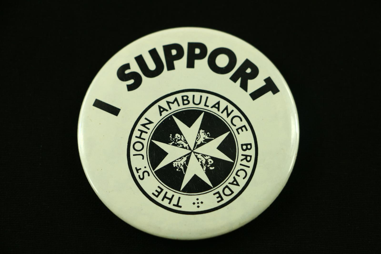 Round, black and white metal badge with plastic coating. The badge has a white background. At the top is black text 'I SUPPORT' arranged in an arch shape. Under the text is the circular St John Ambulance logo in black. It has the text 'THE ST JOHN AMBULANCE BRIGADE' around the circle with a star shape in the centre made from 4 pointed triangular shapes. The back of the badge has a safety pin style clip to attach it to clothing.