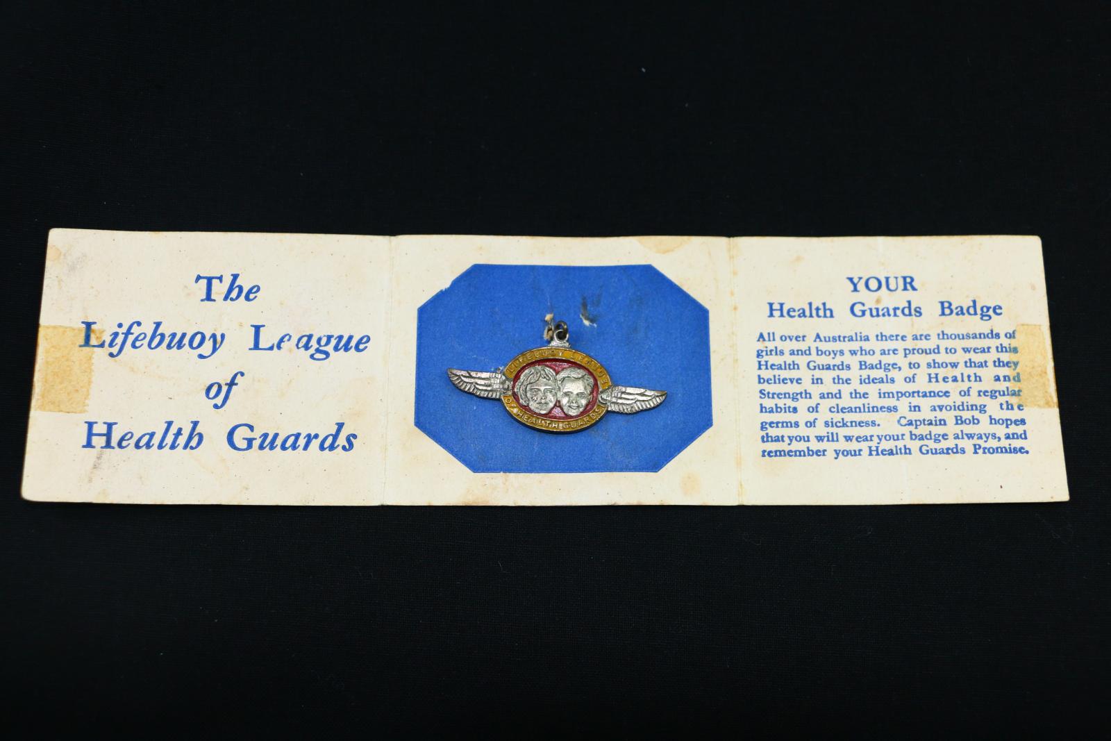 Round, red and yellow, metal, 'Lifebuoy League of Health Guards', metal pin. The pin is held in a blue and white cardboard holder, with text on either side. 