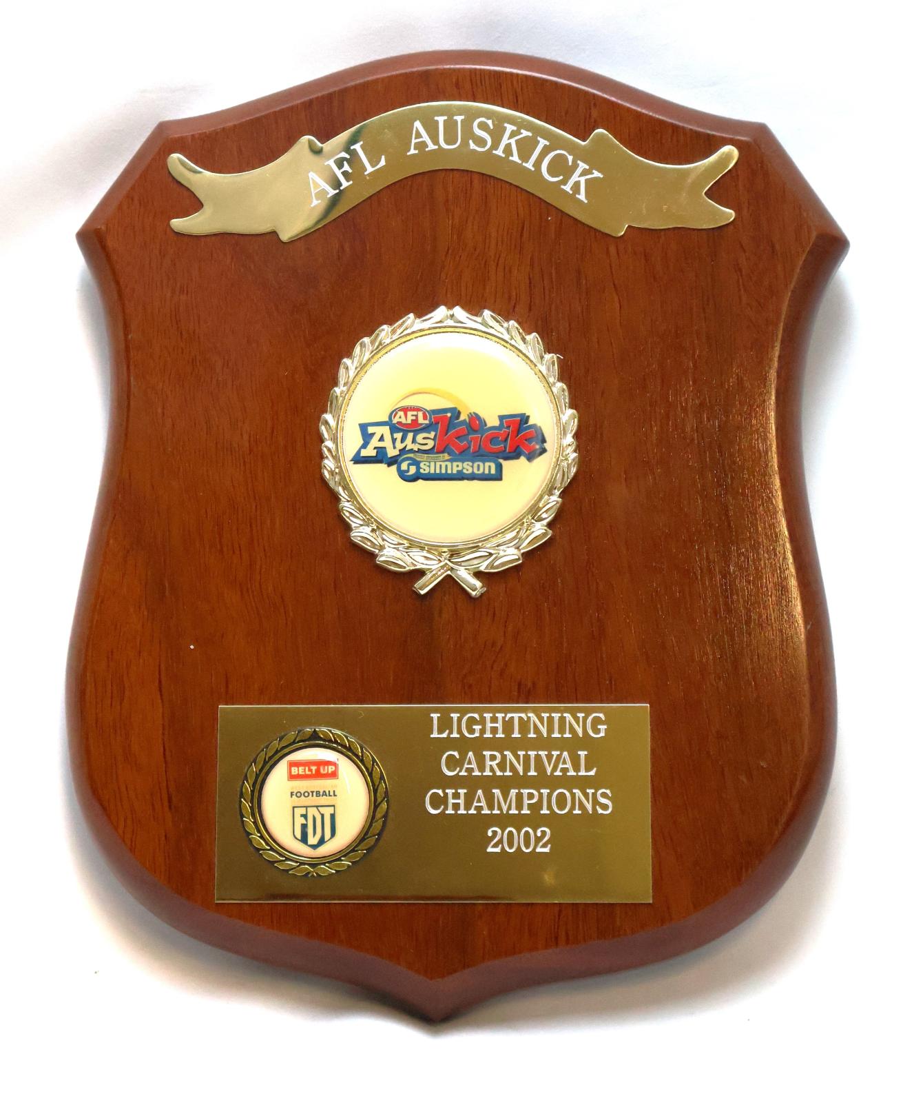 Brown treated and glazed wooden shield shaped plaque with five points. Obverse side has three key features featuring engravings and inscriptions. Along the top is an engraved metal ribbon banner. In the middle there is a raised circular emblem encased in a gilded wreath featuring sponsorship details and AFL Auskick logo. Towards the bottom is a metal rectangle with engraving on the right side and a smaller raised wreath on the left which has extra sponsorship details. 