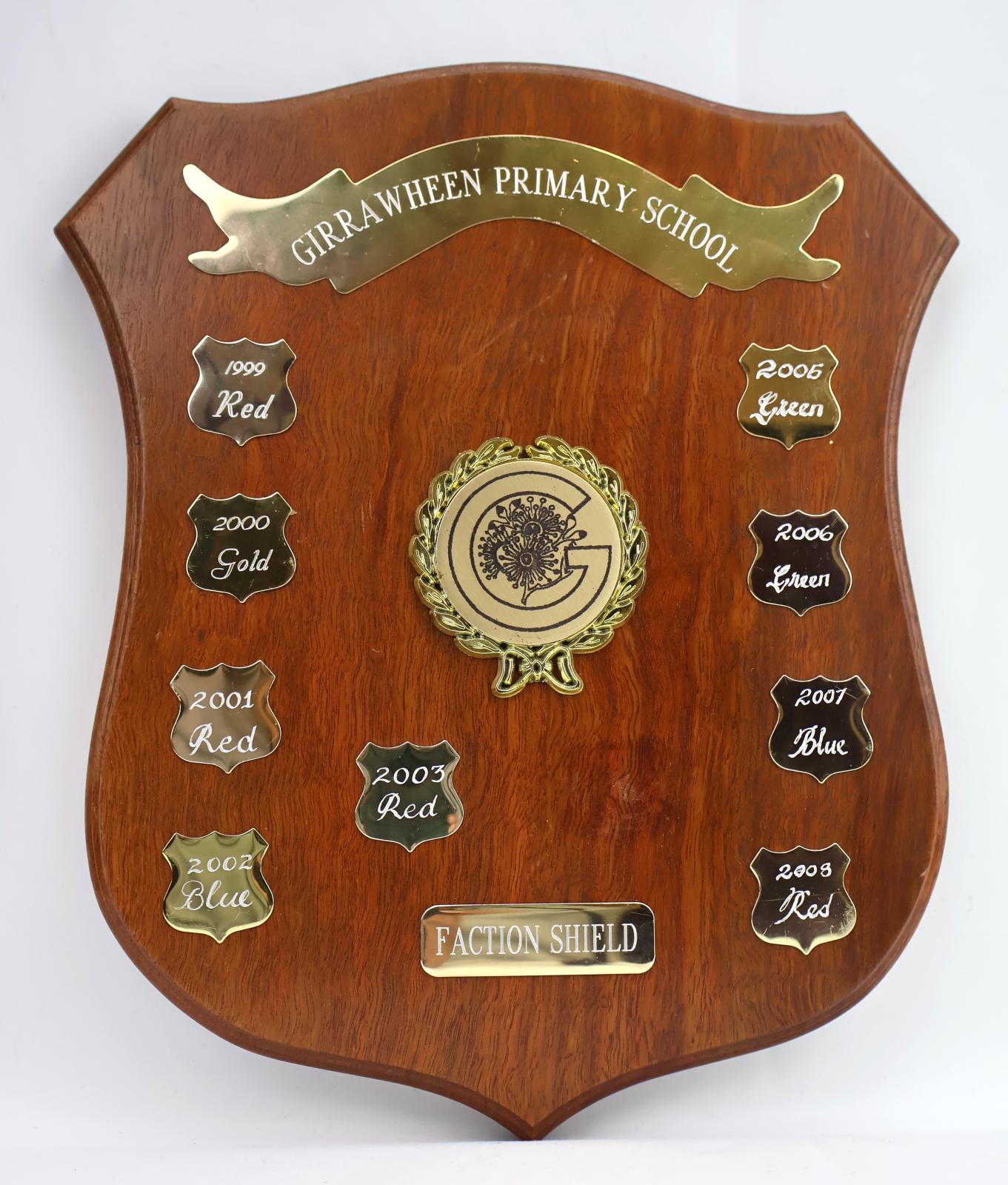 Brown treated wooden shield with four points. The obverse side of the shield consists of nine smaller silver shields with each having six points, with only two of the shields featuring engraving of the year and winning faction team. At the top of the shield there is a raised metal ribbon banner that is held in place by two screws and displays the school’s name. 