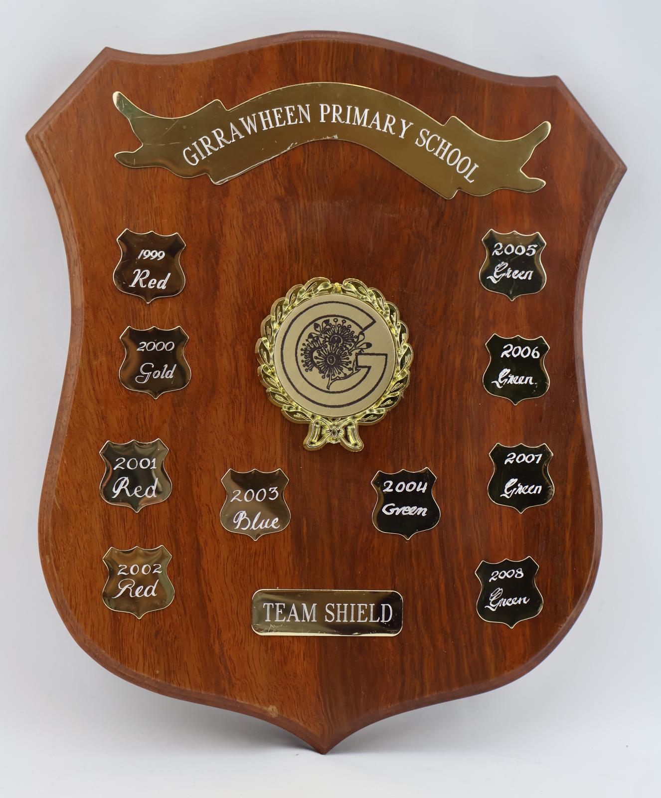 Brown treated wood shield with five points. The obverse side of the shield consists of 10 smaller engraved metal shields featuring team colours and year. At the top of the shield there is a metal ribbon banner. In the middle there is a raised metal wreath with a bow which cocoons around a large letter G and floral emblem. Towards the bottom of the shield is another smaller rounded rectangle that is also engraved.