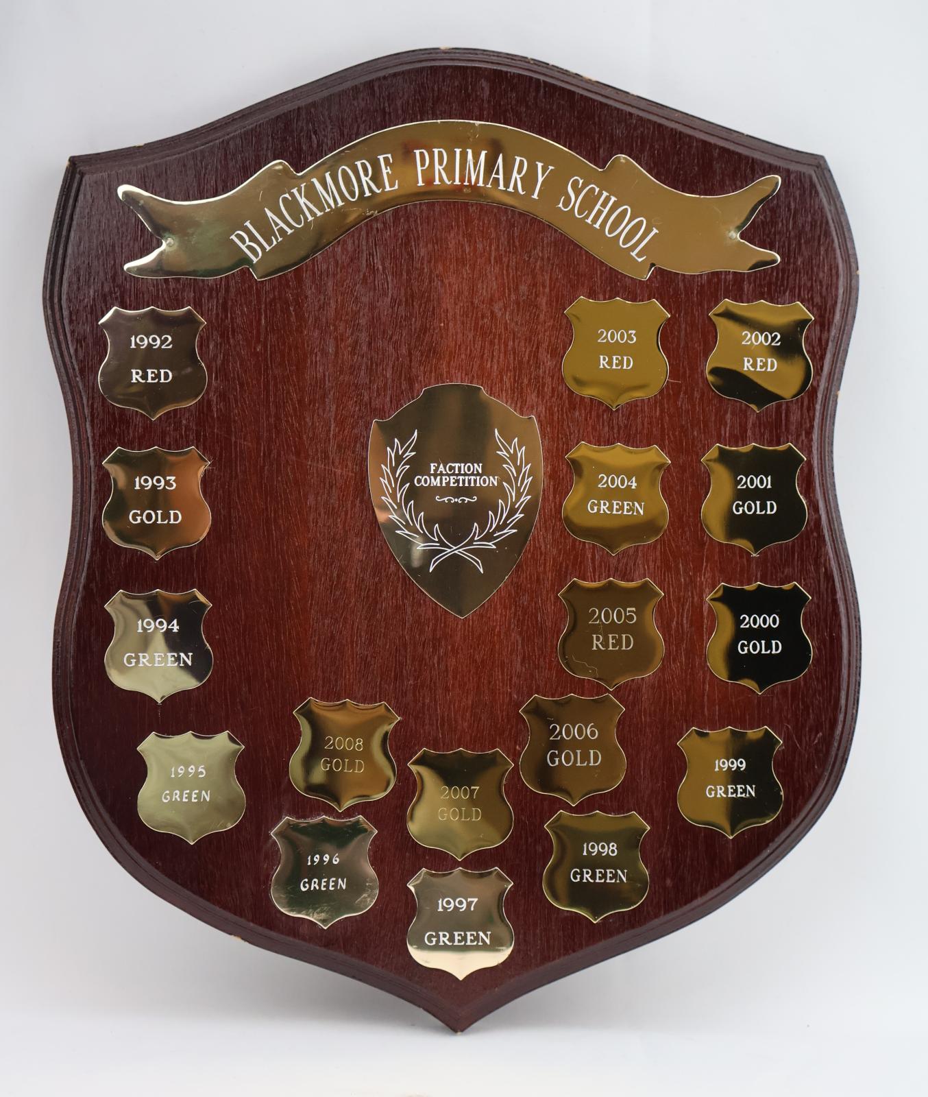 Dark brown treated wooden shield with three points. The obverse side of the shield consists of 17 small shields detailing the year and winning faction colour. At the top of the shield there is a metal ribbon banner engraved with the school’s name. In the centre of the shield is a larger shield of five points that is engraved with a floral emblem and details of the competition. The reverse side of the shield is untreated and has the details of the manufacturer stamped near the top.