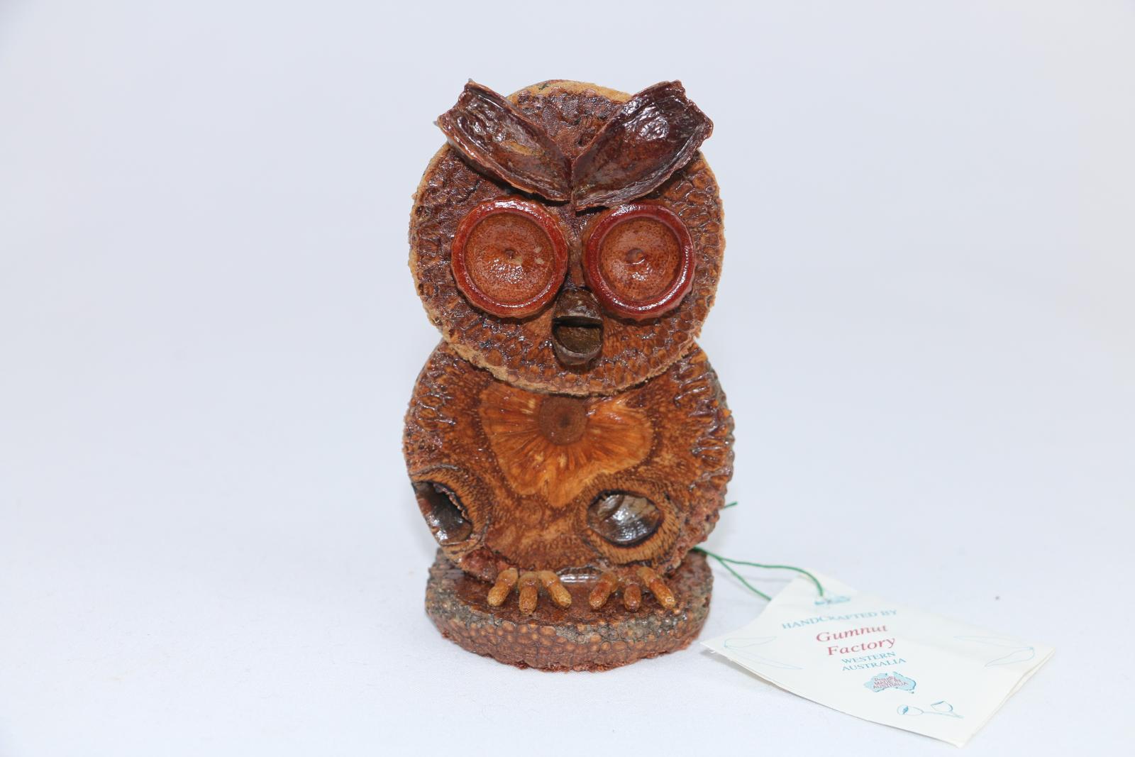 Gumnut owl toothpick holder made of Banksia nut cross cut section glued together as an 'owl'. Seedpods on the 'head' denote eyes, mouth and brow. Feet are also of seed pods. A gumnut is positioned at the rear. The figurine is varnished in clear lacquer. Attached to the feel is a small square paper card.