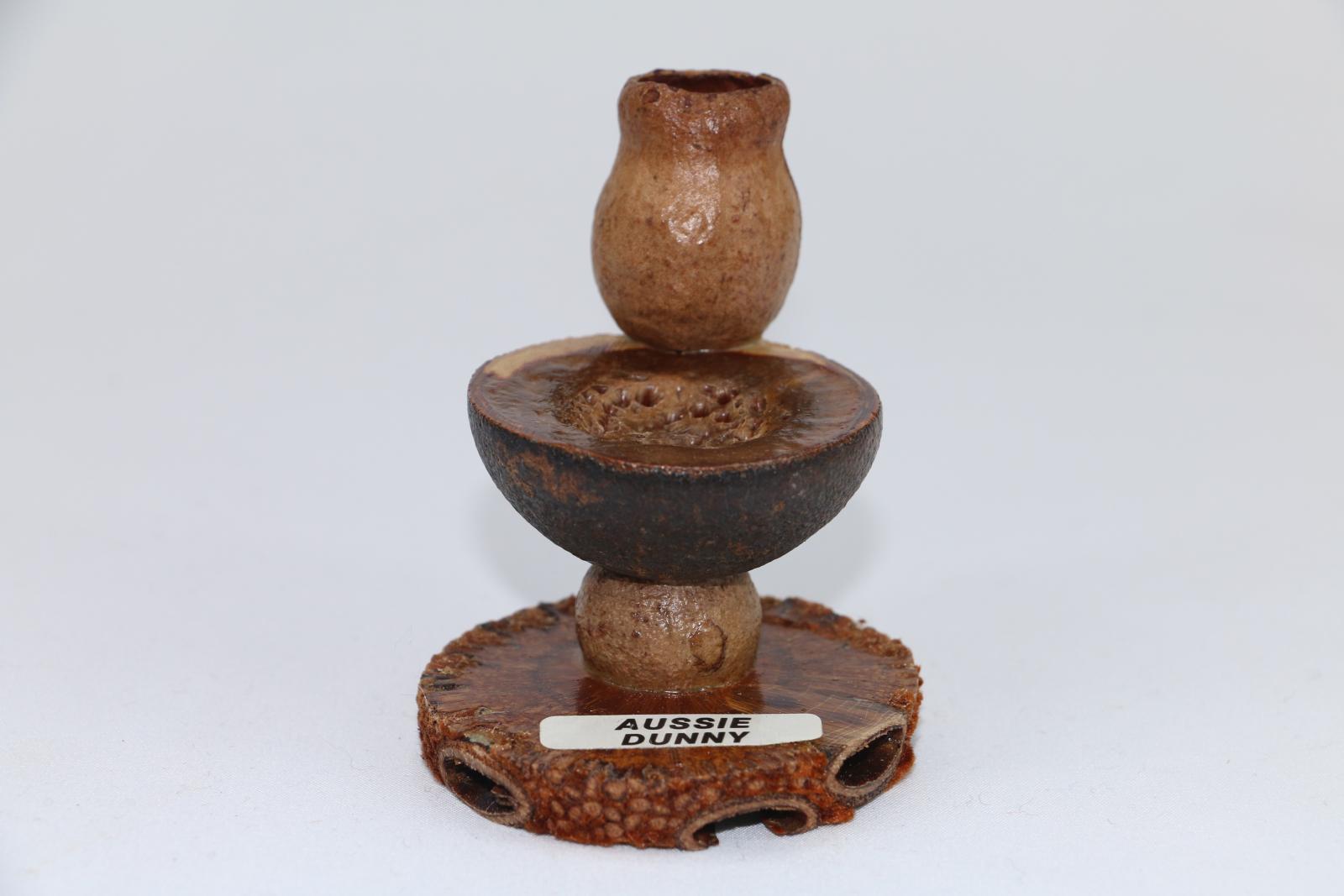 Toilet gumnut figurine made from cross-cut Banksia nut with glued nut supporting a mallee type nut with hollowed-out cross-section depicting toilet bowl with gum nut atop depicting cistern. A label with words 'AUSSIE/ DUNNY' is attached to the Banksia base. The figurine is varnished with clear lacquer.
