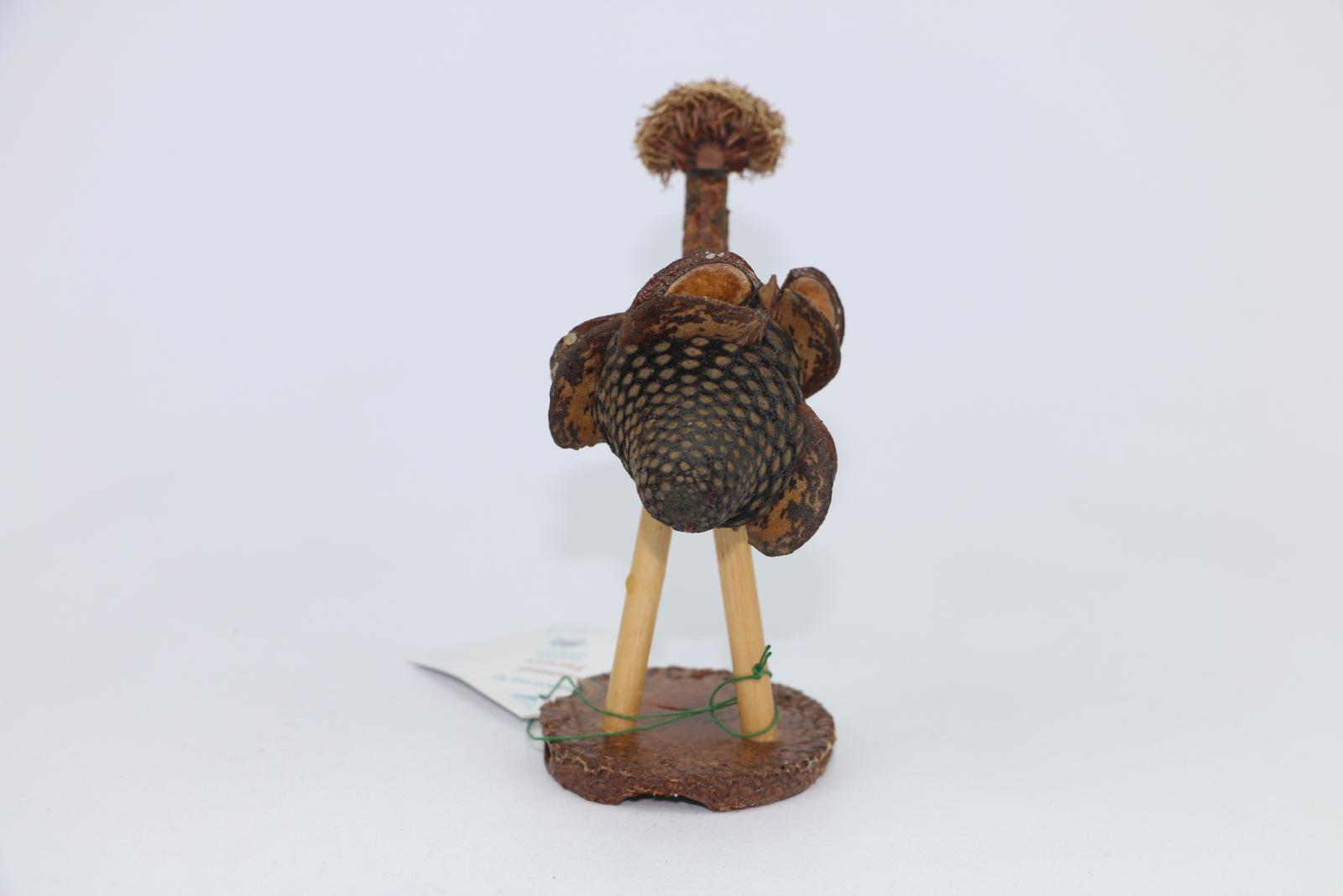 A nut figure in shape of emu is fashioned from Banksia nut for body, stem for neck and hairy nut for head and seed cap for mouth. Stands on two pine 'legs' attached to cross-section of Banksia nut base. Object is coated in a clear varnish. Attached to the feet is a small, square, white paper label.