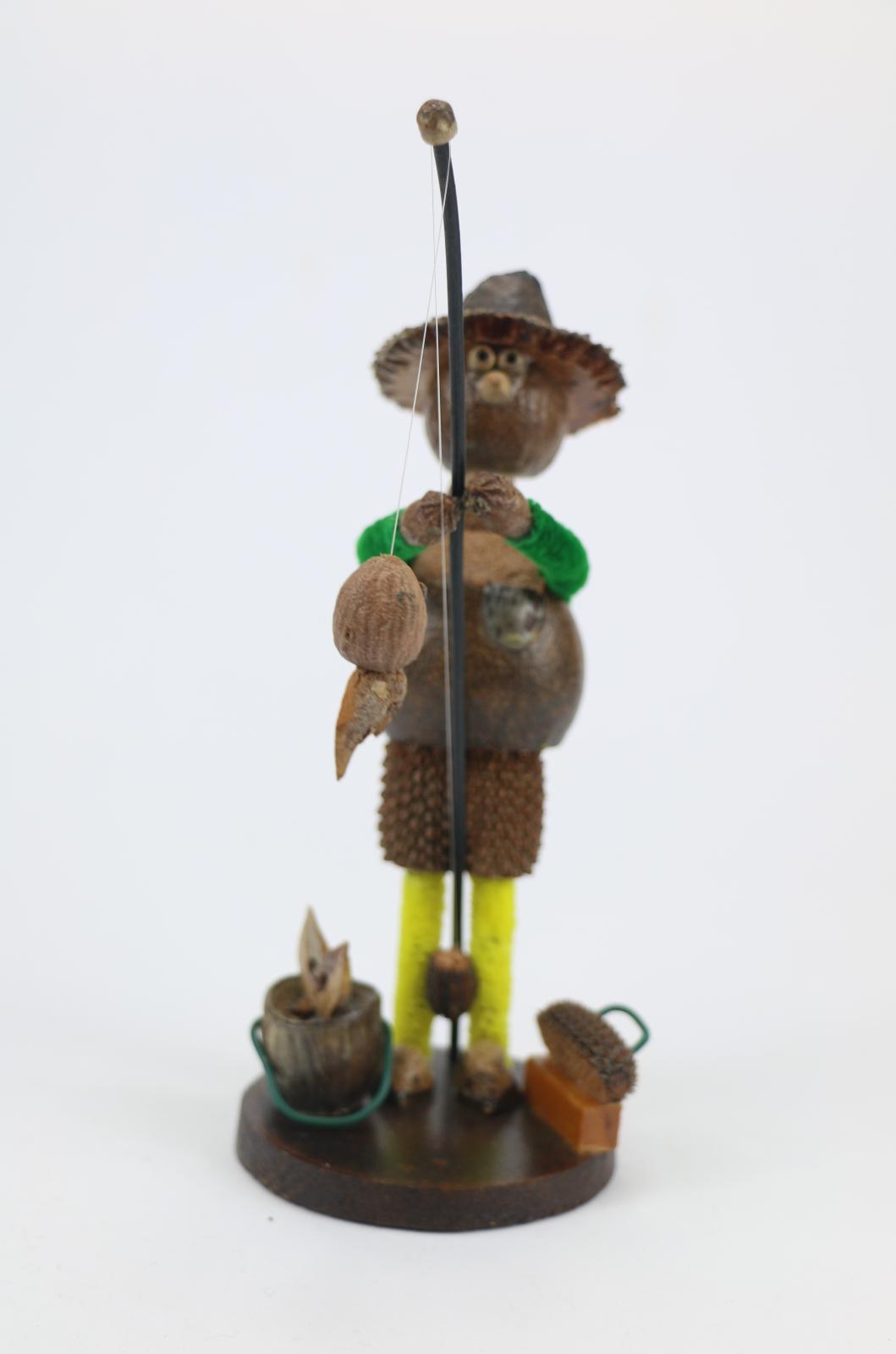 Gum nut figurine fashioned in the style of a fisherman. The figurine is made of gumnuts and has yellow pipe cleaners for legs and green pipe cleaners for arms. The figurine is glued on a cross-cut section of wood that is varnished. The 'fisherman' is holding a length of plastic or nylon 'line' with caught fish on rod. A gumnut stylised as a bucket with plastic handle and a box with plastic handle for bait, hooks etc. is resting on base.