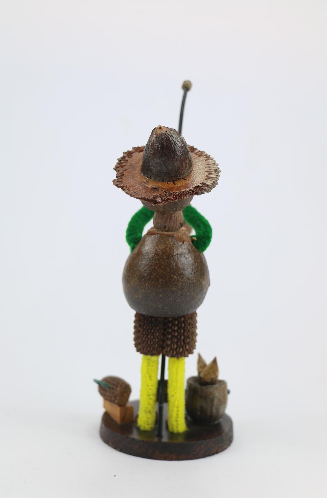 Gum nut figurine fashioned in the style of a fisherman. The figurine is made of gumnuts and has yellow pipe cleaners for legs and green pipe cleaners for arms. The figurine is glued on a cross-cut section of wood that is varnished. The 'fisherman' is holding a length of plastic or nylon 'line' with caught fish on rod. A gumnut stylised as a bucket with plastic handle and a box with plastic handle for bait, hooks etc. is resting on base.