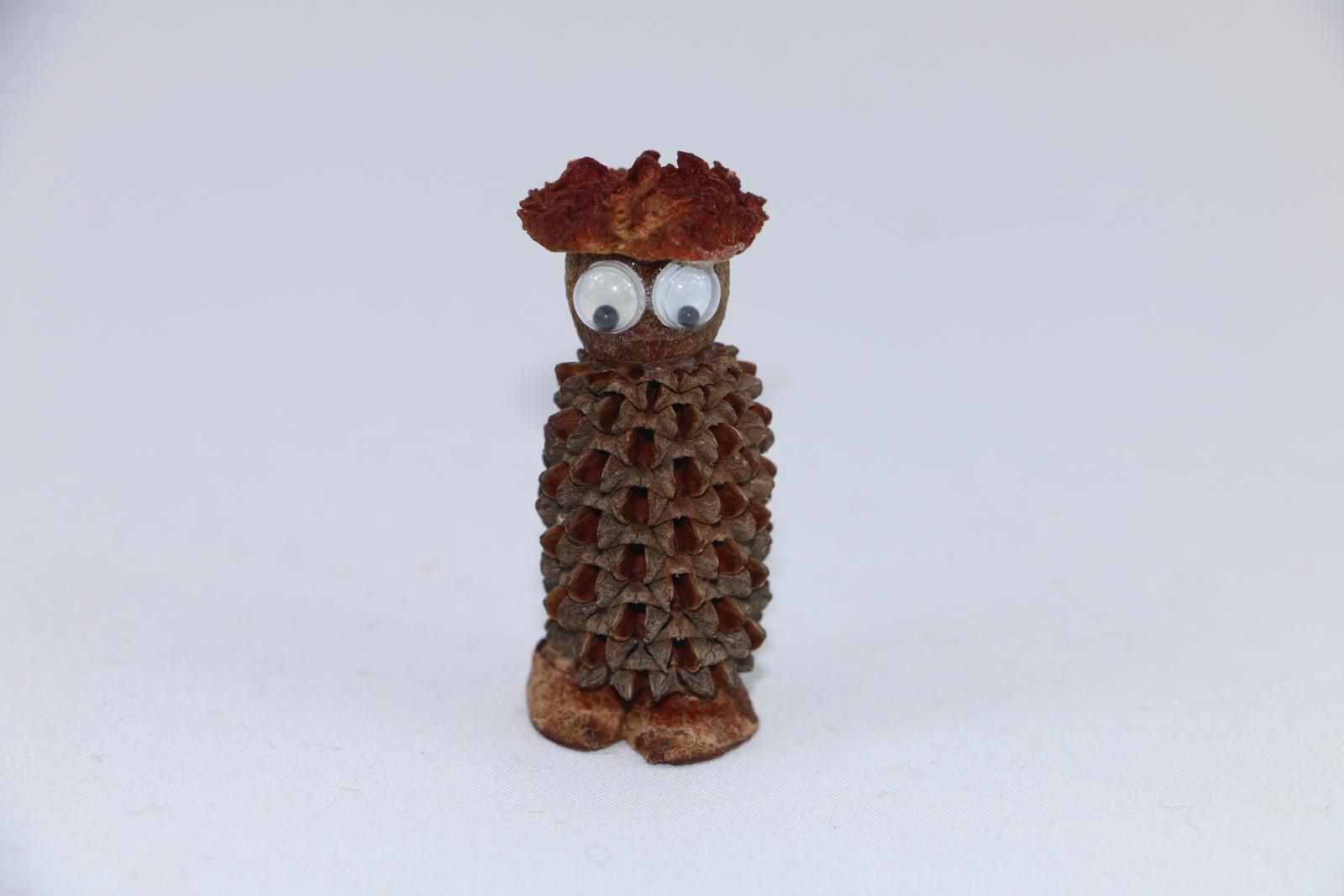 A sheoak nut with a large nut glued on top to represent a head.. Glued to the front of the nut are two large, plastic, goggle eyes. On top of the nut is glued a seed cap to represent a hat. Underneath the sheoak nut are two small split seeds to represent feet.