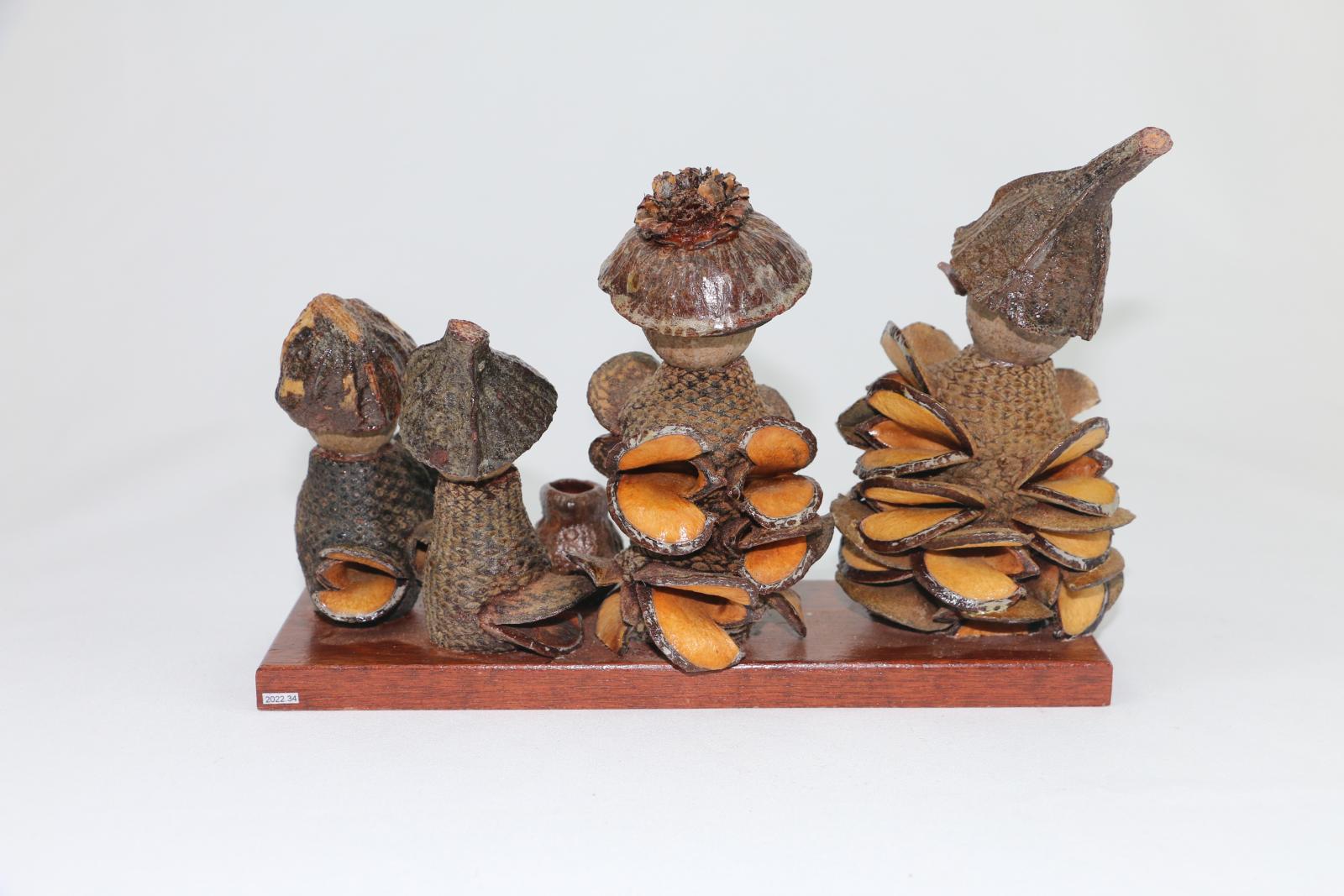 Back View. Four gumnut figures, two adults and two children on a rectangular wooden board with a gumnut in front of figures two and three. Each gumnut figure is made from different parts of Banksia and Eucalyptus trees. Each gumnut is wearing a 'hat' and they all have 'eyes', a 'nose', and a 'body'.