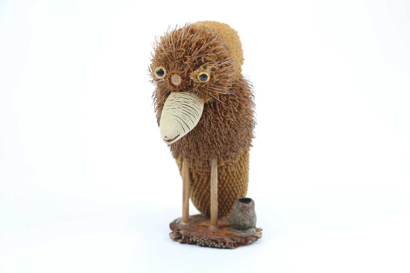 Different plant materials have been used to make a bird figure, there are two plastic eyes attached to the face, the figure is attached to a wooden circular base, there is a smaller gumnut to the right of the figure.