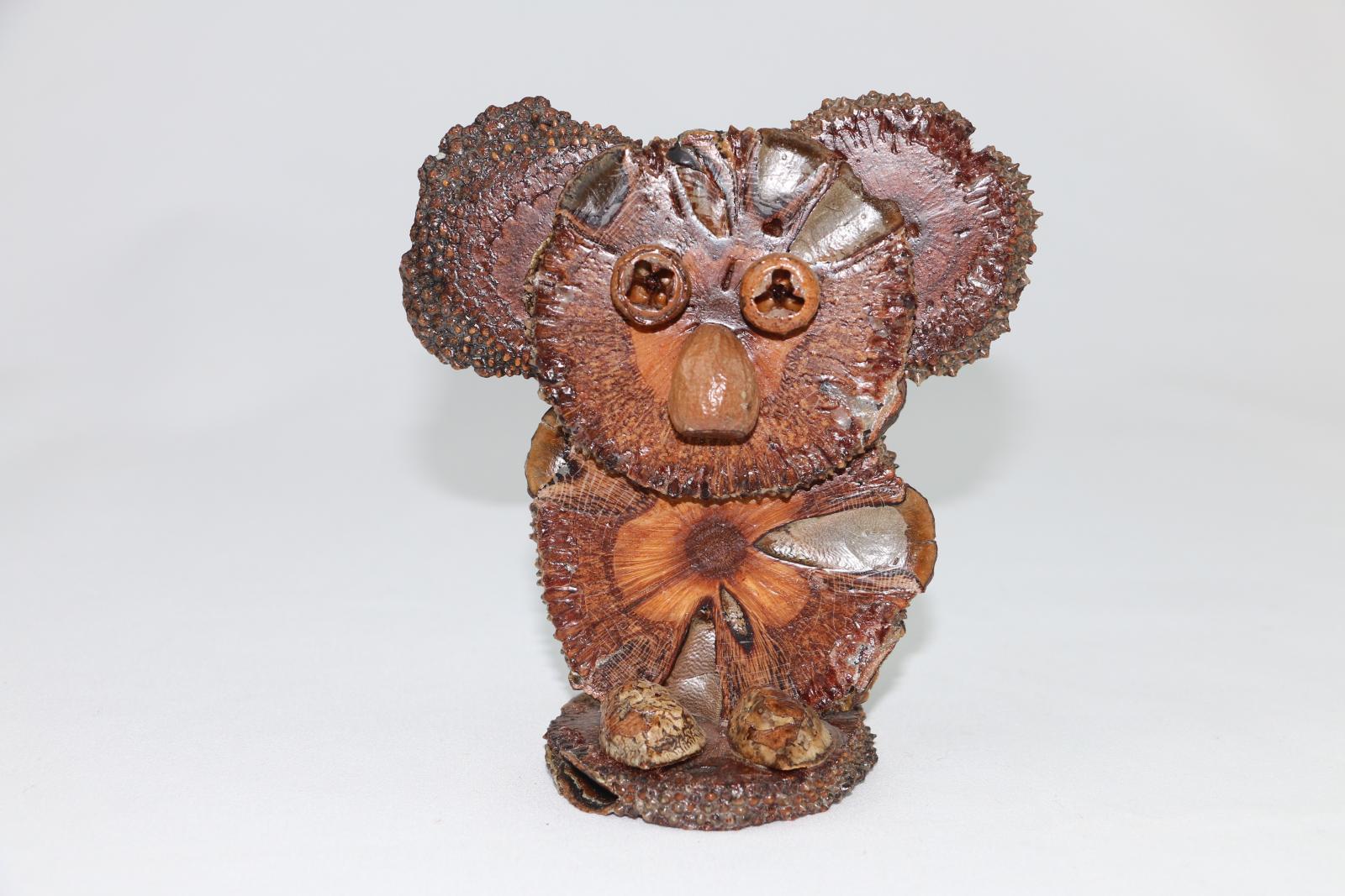 A koala figure made from flat wooden disks and plant material on a circular base. Some of the lacquer has separated from the figure and are forming cracks in them. There is a gumnut at the back of the koala.