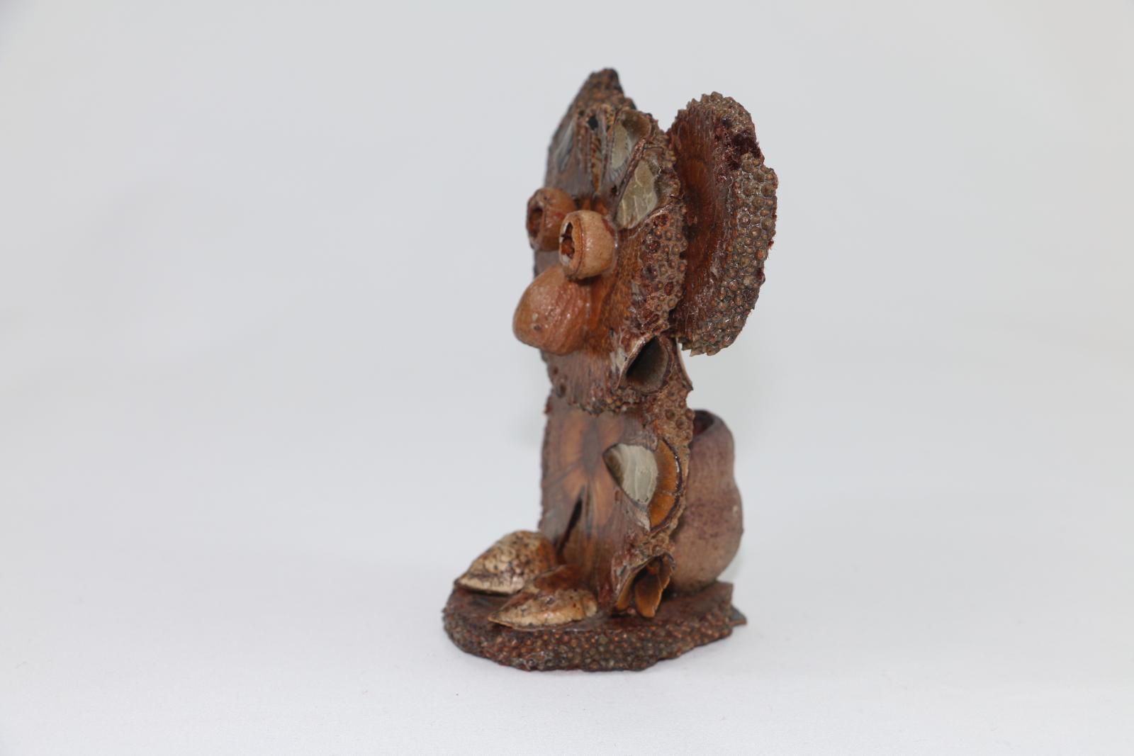 A koala figure made from flat wooden disks and plant material on a circular base. Some of the lacquer has separated from the figure and are forming cracks in them. There is a gumnut at the back of the koala.