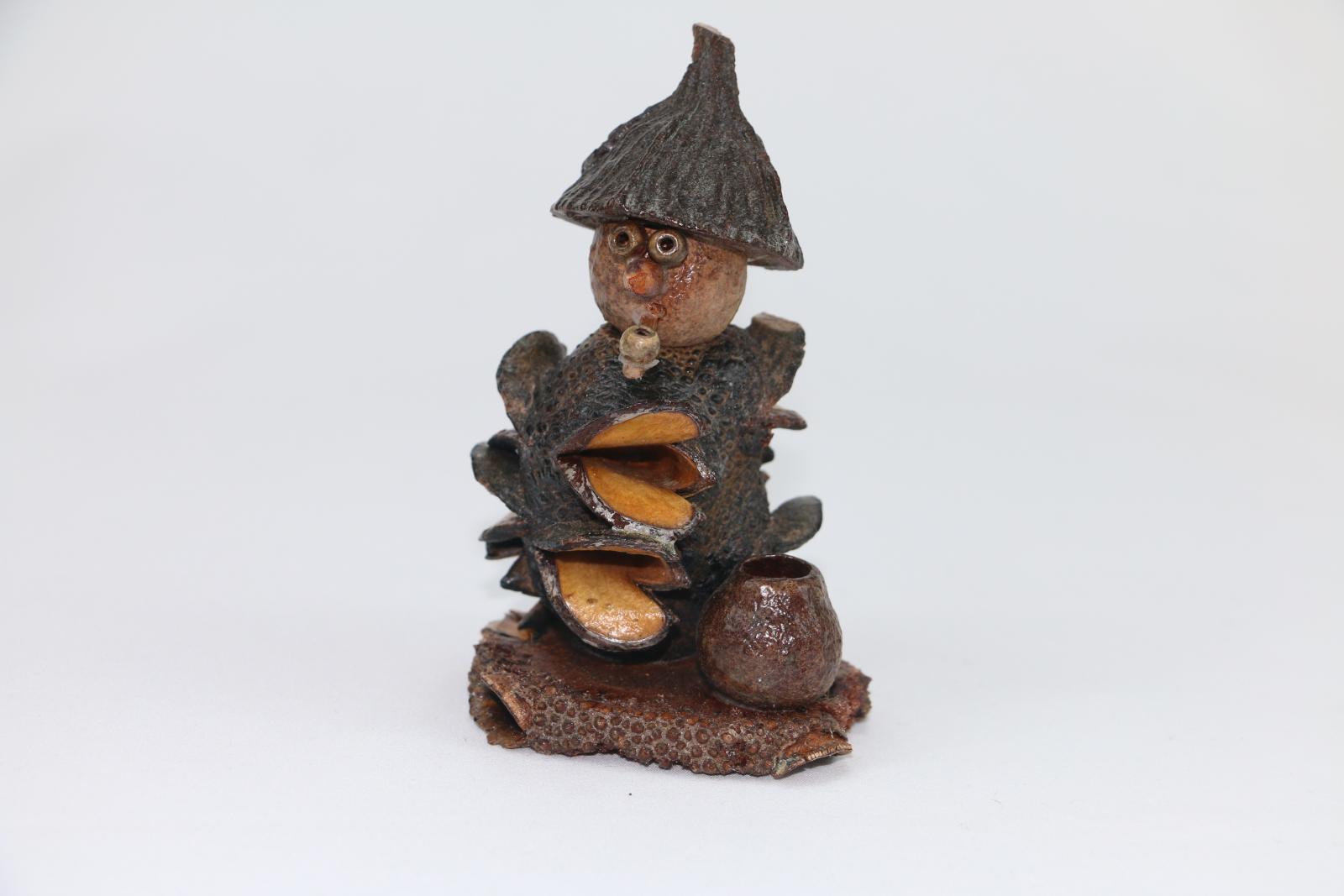 A gumnut figure made from plant material on a circular base made of plant. The figure is smoking a pipe. There is a small gumnut on the left of the figure at the front. 
