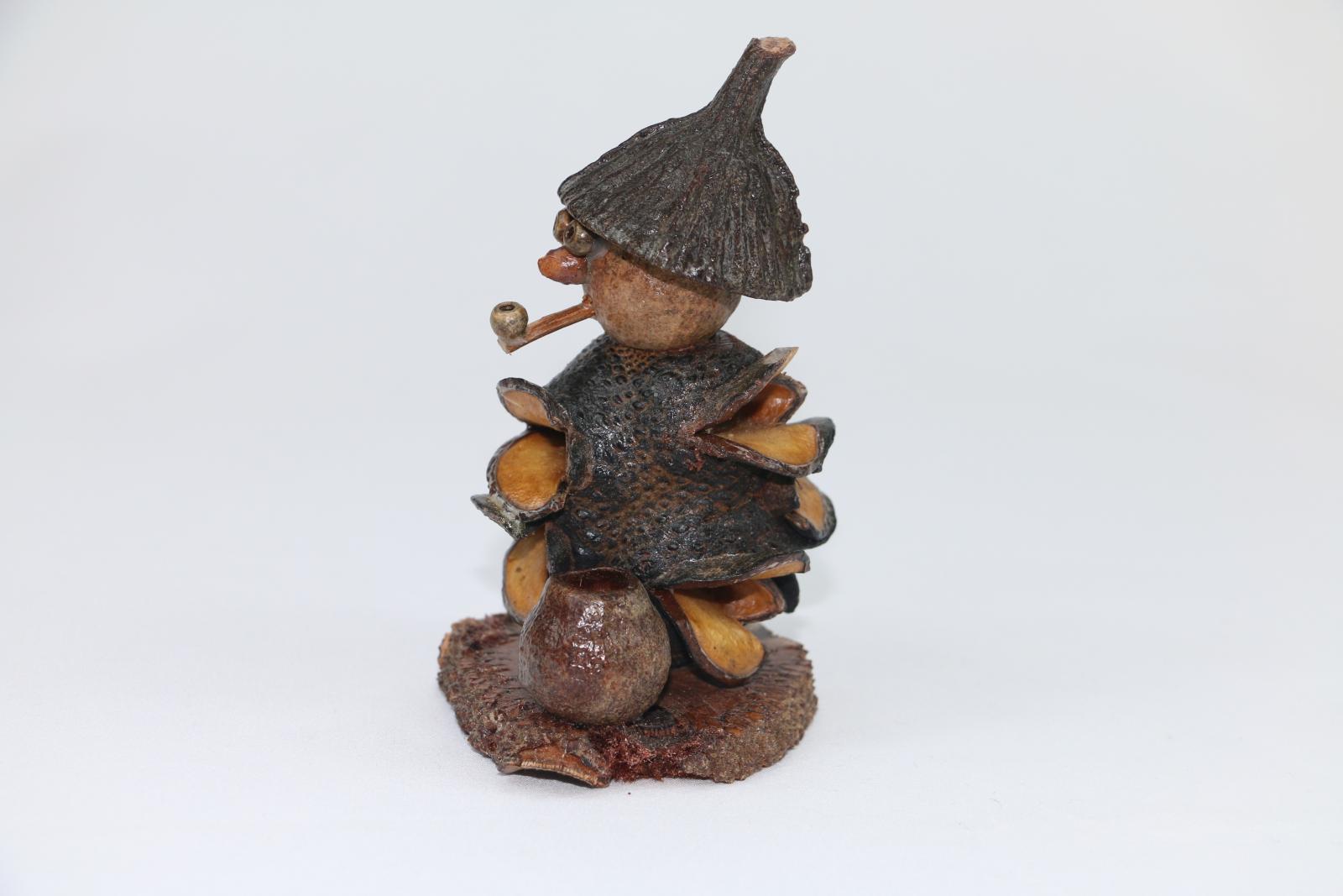 A gumnut figure made from plant material on a circular base made of plant. The figure is smoking a pipe. There is a small gumnut on the left of the figure at the front. 