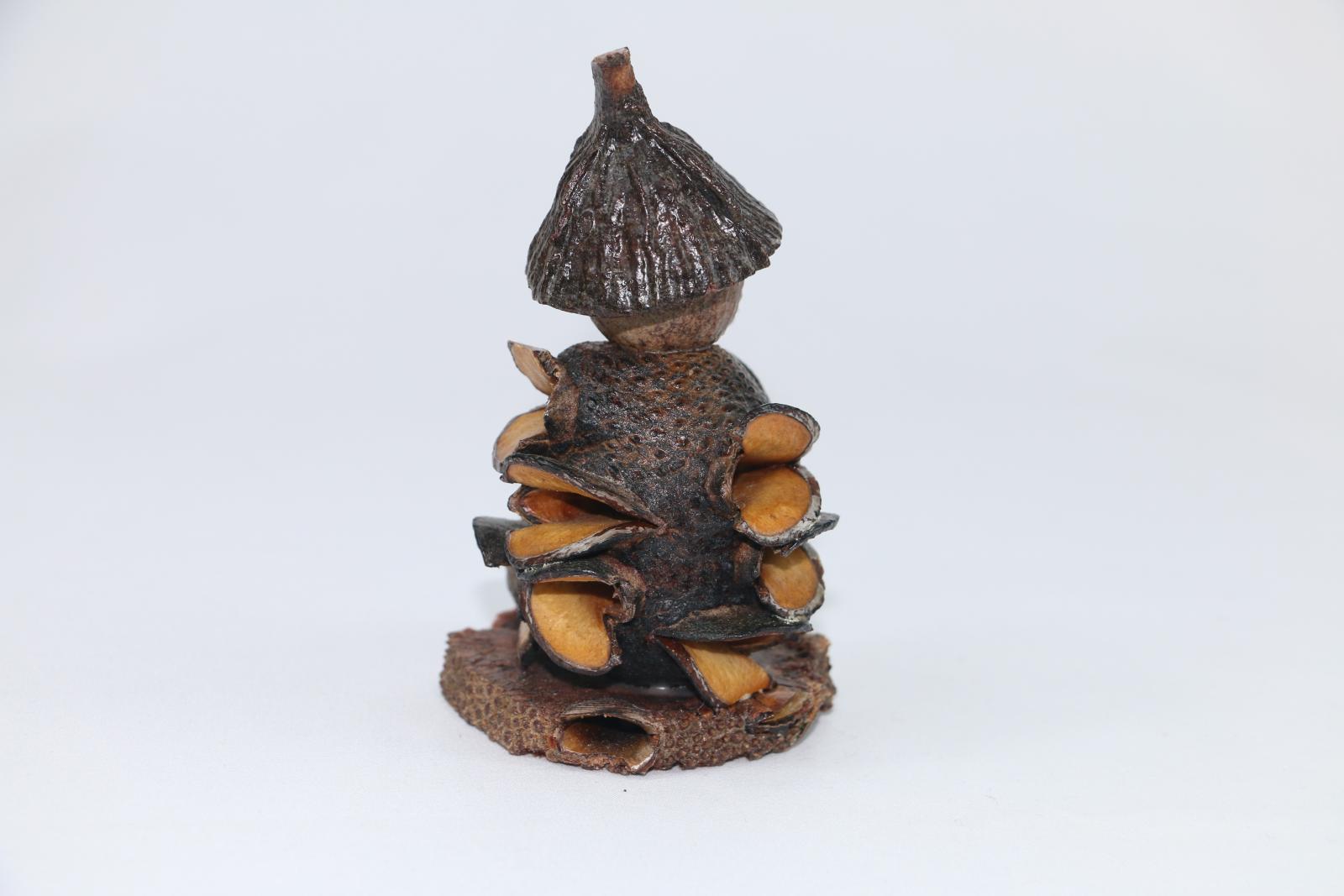 A gumnut figure made from plant material on a circular base made of plant. The figure is smoking a pipe. There is a small gumnut on the left of the figure at the front. 