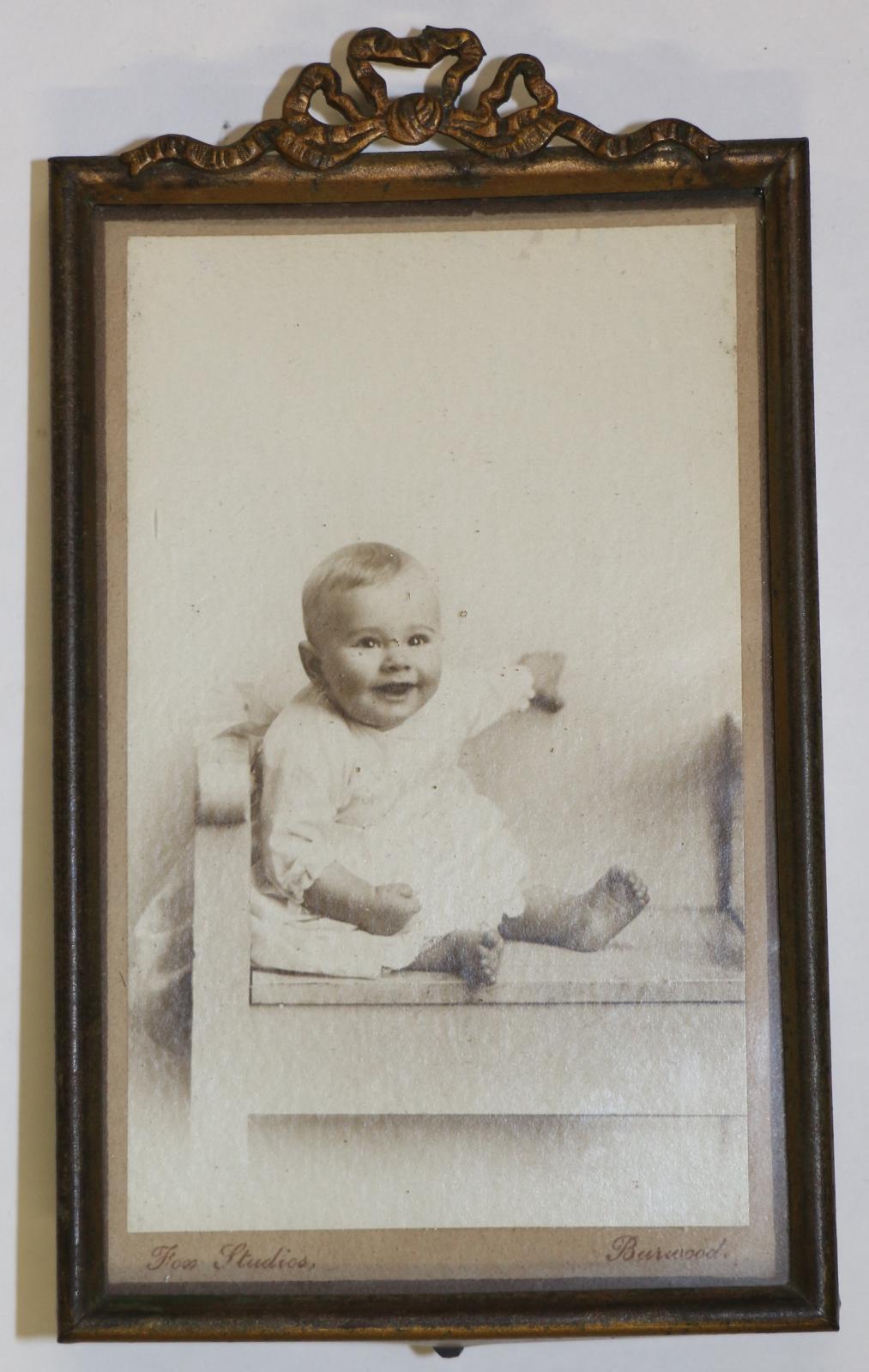 Rectangular bronze metal frame with ecorative bow on the top. Backing made from dark wood, ''MADE IN/ BIRMINGHAM/ ENGLAND" stamped in a circle on hinged stand. The glass is covering a black and white photograph of a baby sitting on a chair.