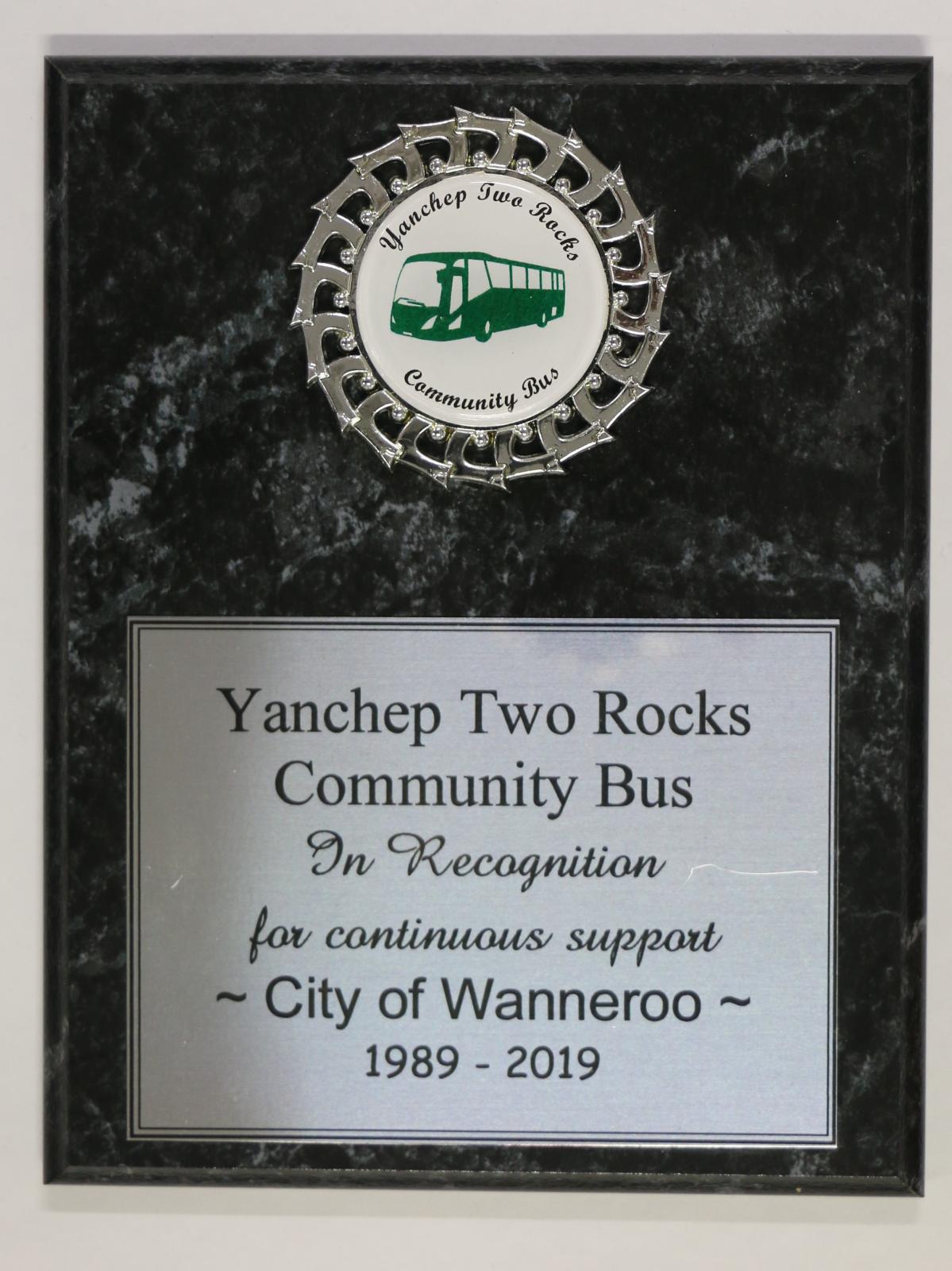 A wooden backing with a black marble look finish. A silver with black border plaque, has minor scratches, which says "Yanchep Two Rocks/ Community Bus/ In Recognition/ For continuous support/ ~City of Wanneroo~/ 1989-2019 Badge - Yanchep Two Rocks/ Community Bus". The wood backing has a small chip on the bottom RHS. A circular stain on the top LHS close to the badge. The badge is circular and made from plastic and acrylic. Small damage to plastic badge at top centre. 