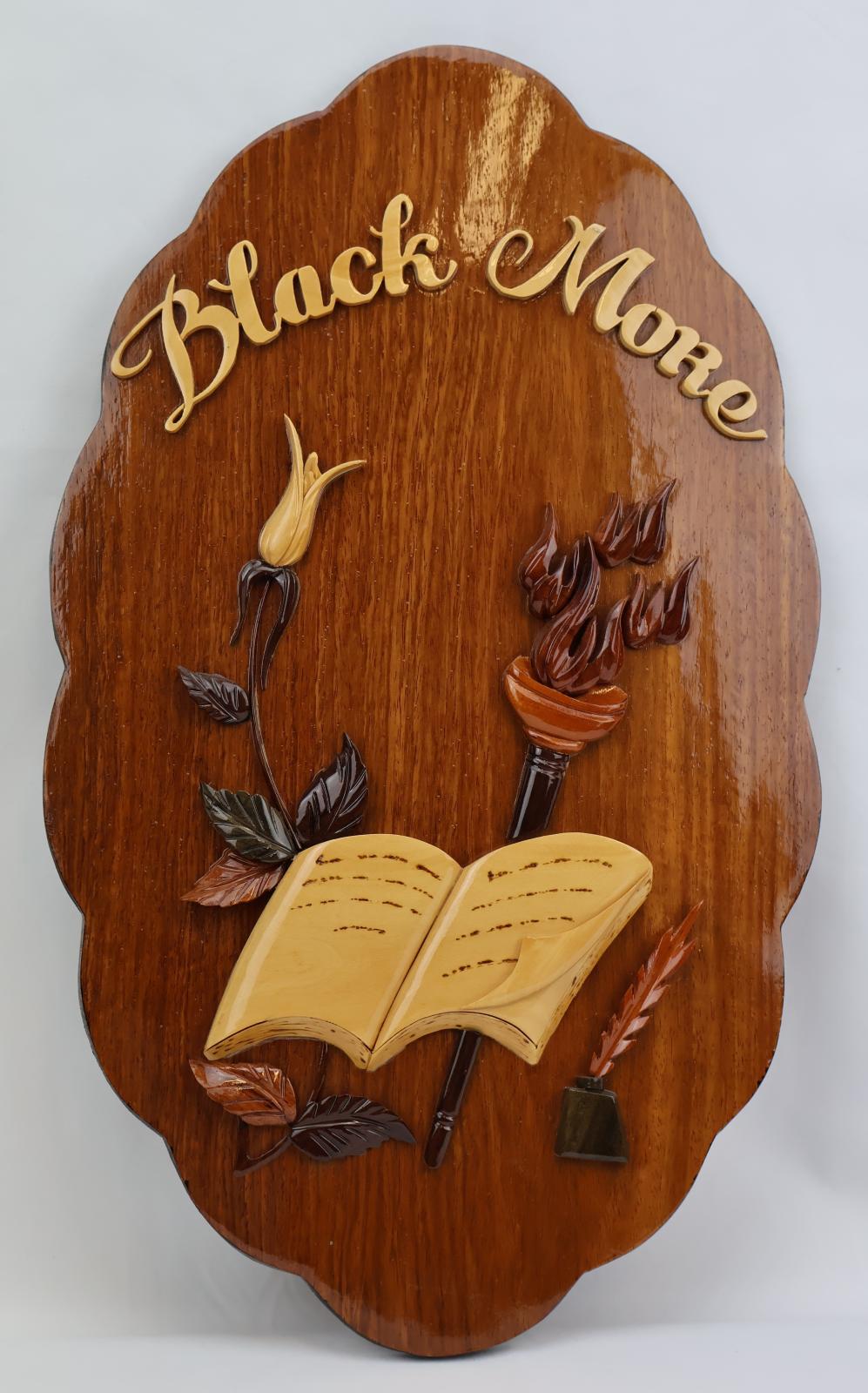 Oval plaque with scalloped edges, decorated with wooden pieces depicting a flower, a torch and an open book. Black More is written across the top with lighter wood pieces.