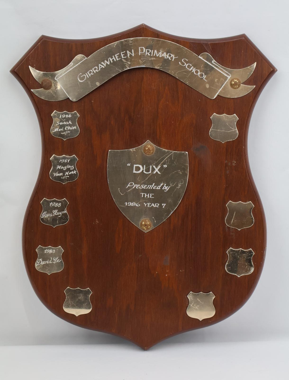 Brown wooden shield shaped plaque. Metal banner shaped attachment across the top with the school’s name. In the centre of the shield is a smaller metal shield made from metal with ‘Dux Presented by THE 1986 YEAR 7’. On the left of the shield is 5 engraved metal shields.