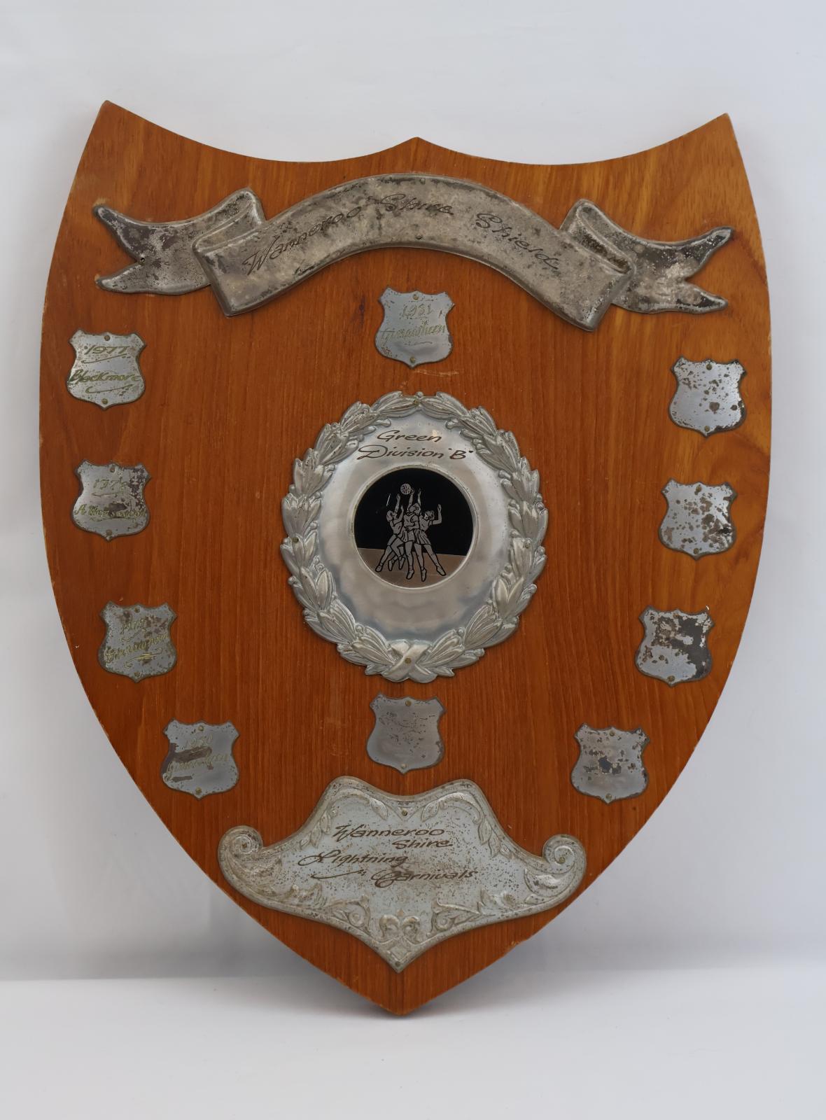 A brown wooden shield shaped plaque. There is a raised silver metal banner with text in the middle. In the middle of the plaque is a circular badge with a wreath around the edge, at the top is text and the centre has an image of three women reaching for a ball. At the bottom of the plaque is a stylized ornate shield shape which has a decorative edge and text in the centre. On the left side of the plaque on top and bottom of the centre circular badge. 