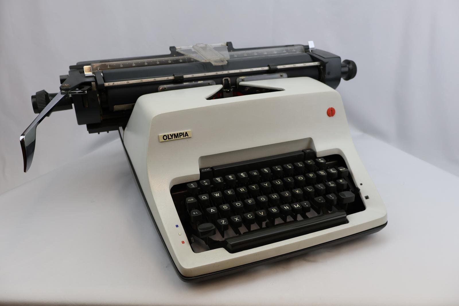 White front with grey keys. A grey carriage with clear plastic paper support. On the carriage is a silver metal line spacer lever. Rectangular shape, four feet on the bottom corners. Raised area in the middle same level as carriage. 