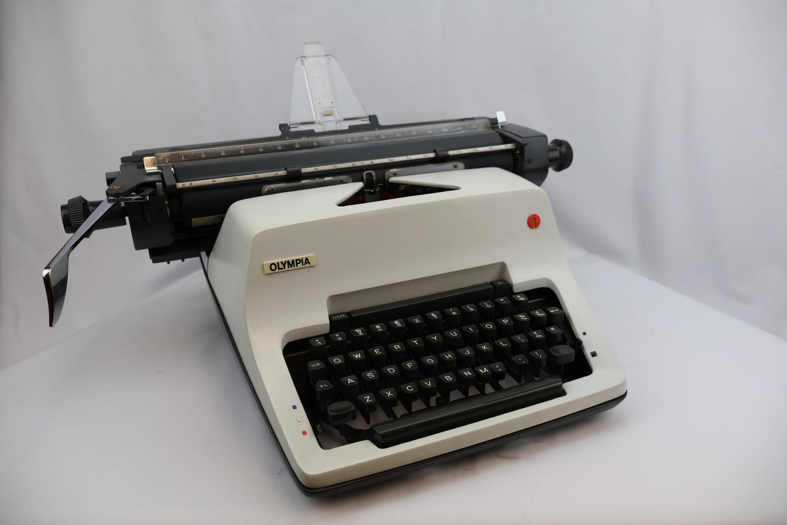 White front with grey keys. A grey carriage with clear plastic paper support. On the carriage is a silver metal line spacer lever. Rectangular shape, four feet on the bottom corners. Raised area in the middle same level as carriage. The front tapers down at an angle for the type keys.