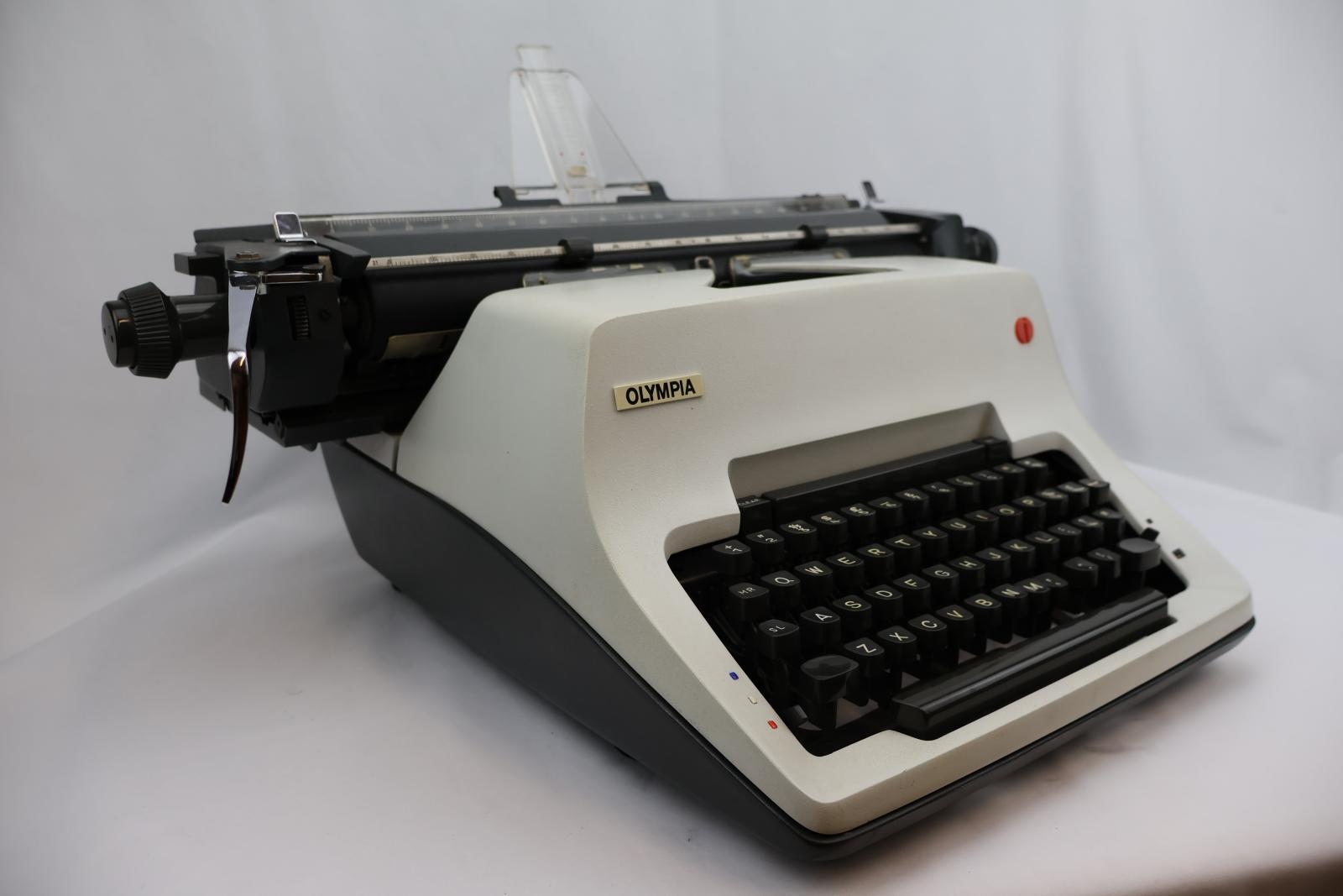 White front with grey keys. A grey carriage with clear plastic paper support. On the carriage is a silver metal line spacer lever. Rectangular shape, four feet on the bottom corners. Raised area in the middle same level as carriage. The front tapers down at an angle for the type keys.
