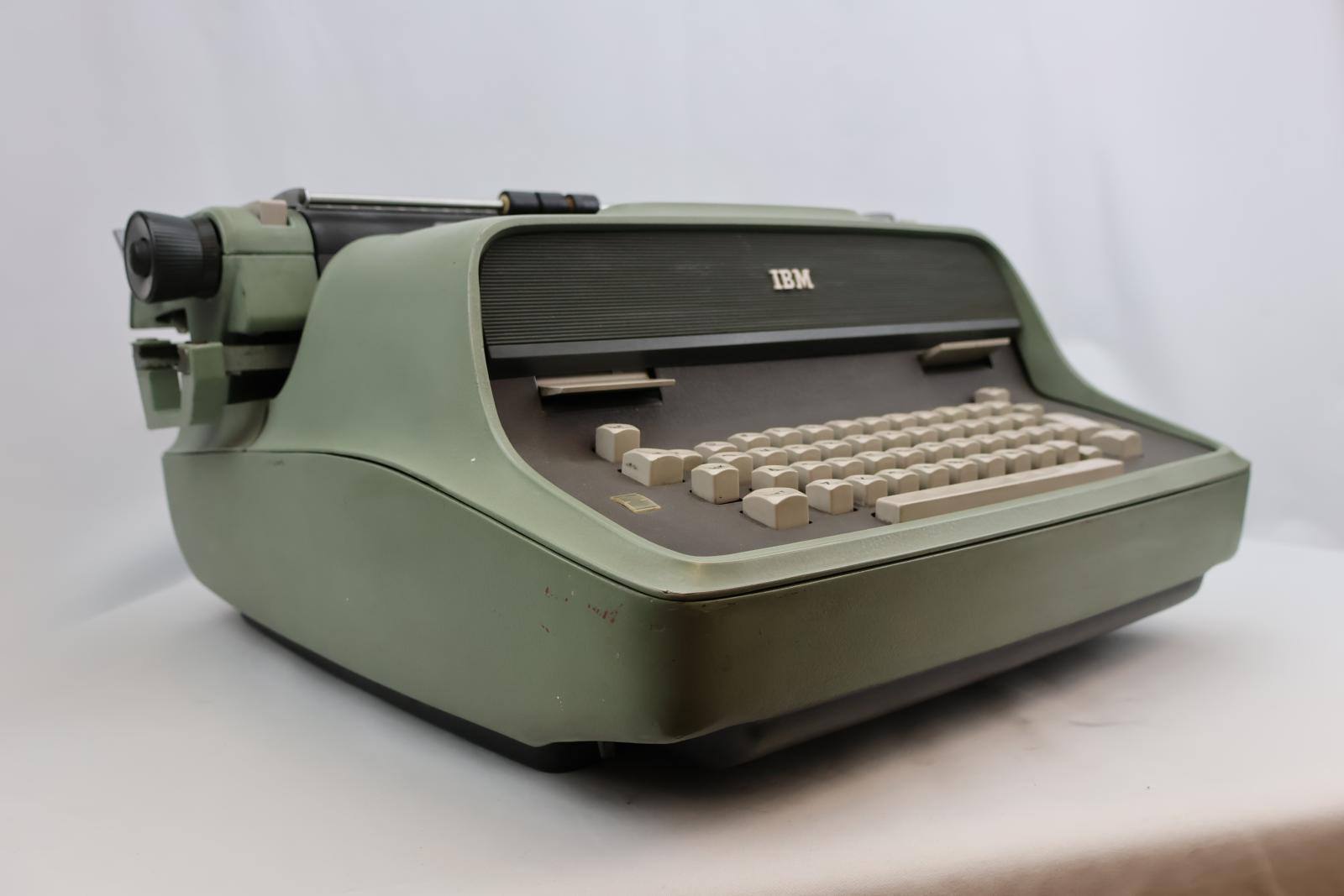 Green electric typewriter, keyboard face is grey with cream keys. Three buttons on top of the keyboard, ribbon position, TAB clear and TAB set. Paper bail and holder black with measurements across the top. The carriage is also green with a black dial at each end and there are also levers and buttons on the carriage. 