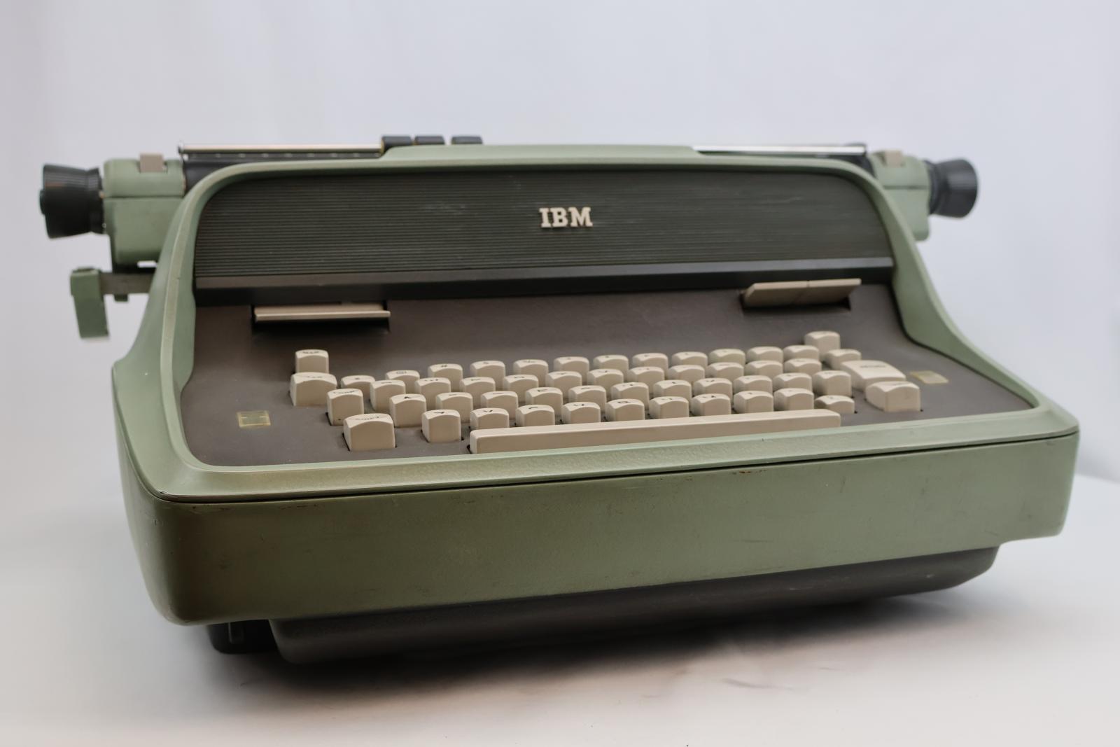 Green electric typewriter, keyboard face is grey with cream keys. Three buttons on top of the keyboard, ribbon position, TAB clear and TAB set. Paper bail and holder black with measurements across the top. The carriage is also green with a black dial at each end and there are also levers and buttons on the carriage. 