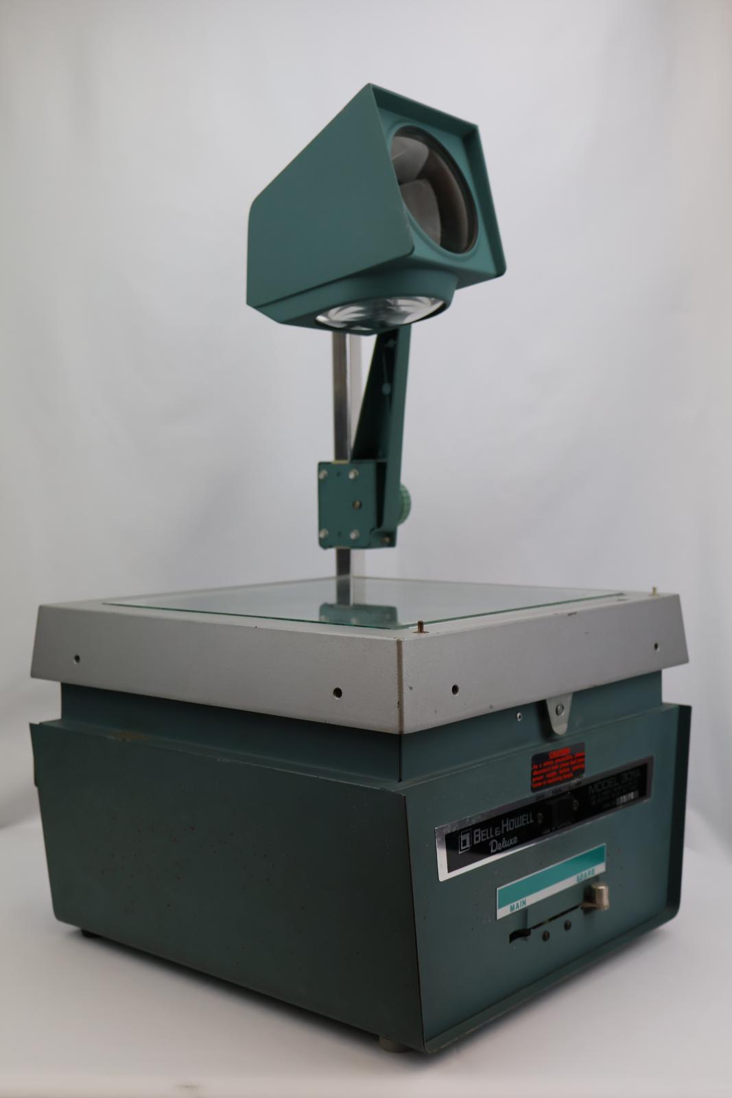 Square  box, green and silver with glass on top. Vent and electrical cable at the back. Silver square pole from back corner with green arm attached, arm has dial to move arm up and down. The top of the green arm has a square with two circular glass lenses. The main body opens at the glass/ silver top. Inside there are wires, a light globe, and a fan.