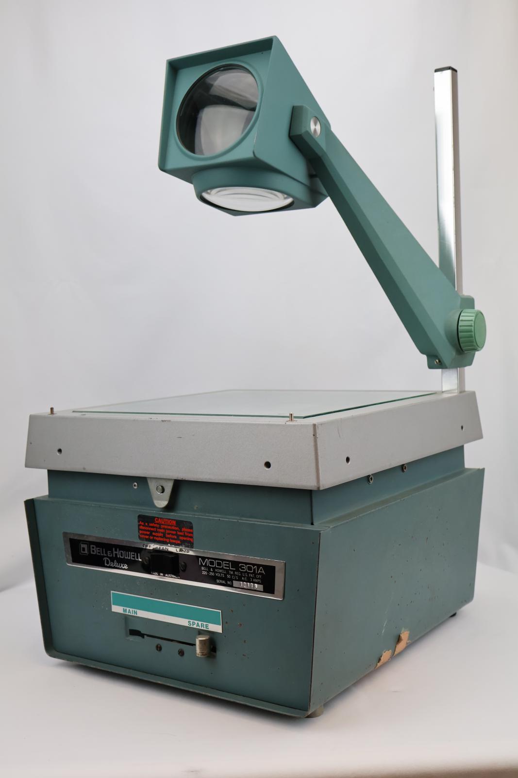 Square  box, green and silver with glass on top. Vent and electrical cable at the back. Silver square pole from back corner with green arm attached, arm has dial to move arm up and down. The top of the green arm has a square with two circular glass lenses. The main body opens at the glass/ silver top. Inside there are wires, a light globe, and a fan.