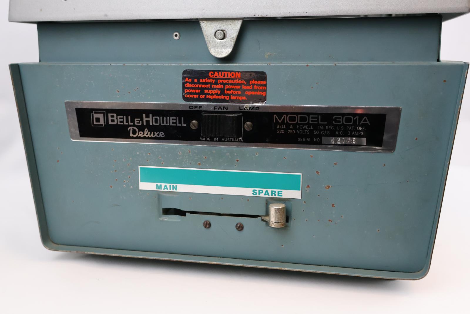 Square  box, green and silver with glass on top. Vent and electrical cable at the back. Silver square pole from back corner with green arm attached, arm has dial to move arm up and down. The top of the green arm has a square with two circular glass lenses. The main body opens at the glass/ silver top. Inside there are wires, a light globe, and a fan.