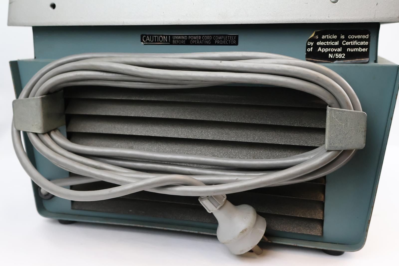 Square  box, green and silver with glass on top. Vent and electrical cable at the back. Silver square pole from back corner with green arm attached, arm has dial to move arm up and down. The top of the green arm has a square with two circular glass lenses. The main body opens at the glass/ silver top. Inside there are wires, a light globe, and a fan.