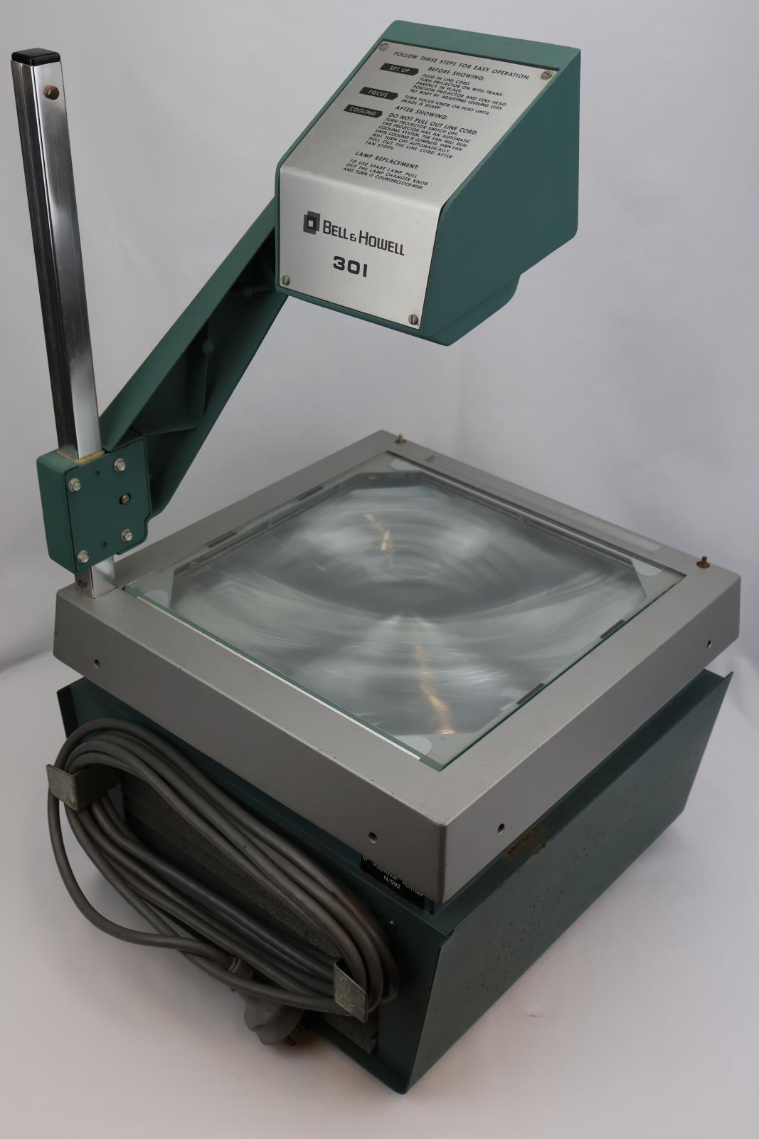 Square  box, green and silver with glass on top. Vent and electrical cable at the back. Silver square pole from back corner with green arm attached, arm has dial to move arm up and down. The top of the green arm has a square with two circular glass lenses. The main body opens at the glass/ silver top. Inside there are wires, a light globe, and a fan.