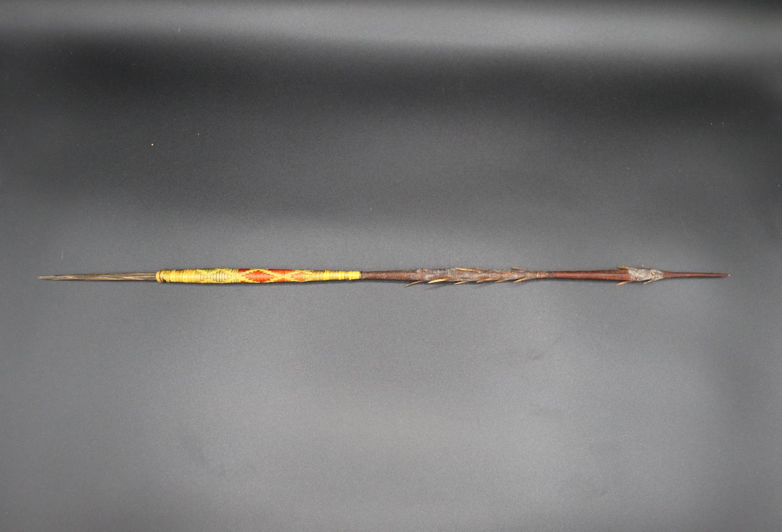 A barbed spear, approx. 45cm long, with a yellow and red patterned grip.