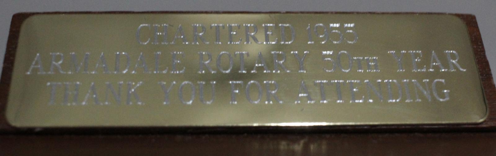 An engraved, gold-coloured metal plate, reading [CHARTERED 1955 ARMADALE ROTARY 50TH YEAR THANK YOU FOR ATTENDING]