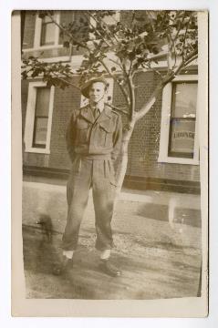 Black and white photo of a male in Australian Army uniform, wearing a slouch hat, standing upright with hands behind back, brick building in background