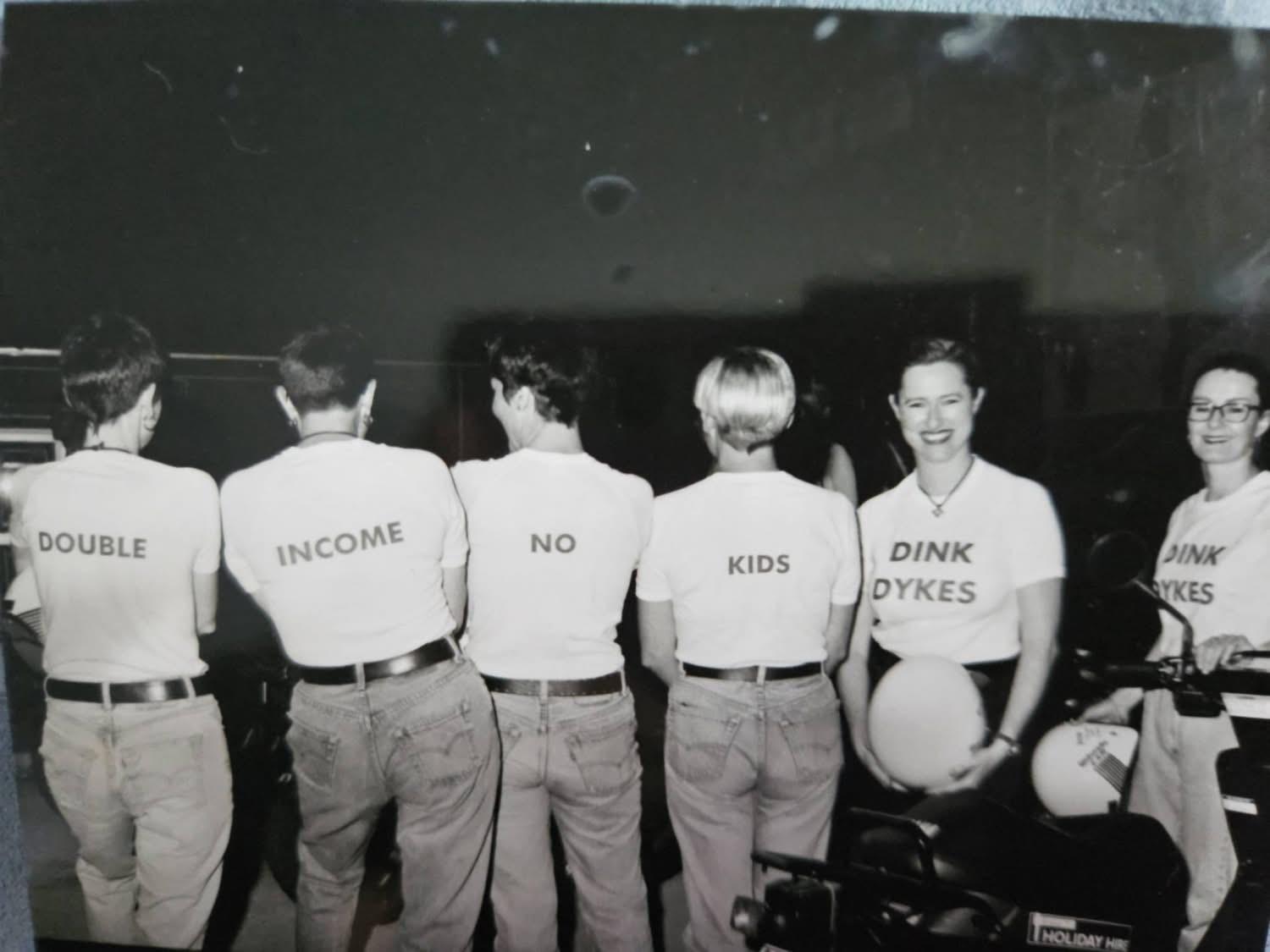 black and white photo of six people wearing white tshirts with writing on them saying DOUBLE INCOME NO KIDS DINK DYKES DINK DYKES