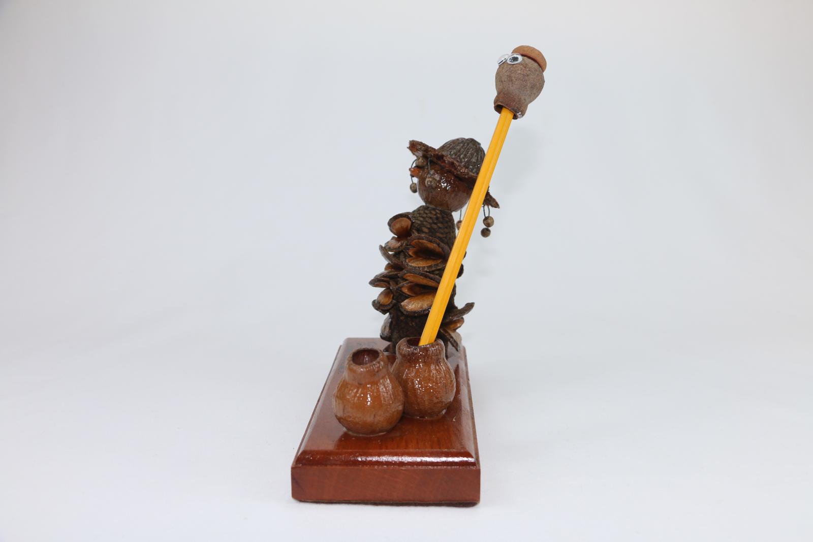 Brown, gum nut figurine sitting on a thick, rectangular wooden stand. A gum nut figure stylised as a swagman serves as an ink or pencil stand. The stylized swagman has a Banksia nut body and gumnut head with another nut as a hat with small beads hanging from it. The wooden base is lacquered with words engraved on the surface 'Compliments of/GUMNUT FACTORY/WANNEROO. W.A.' Two gum nuts on the right of the stand are glued in place and function as a pencil holder. 