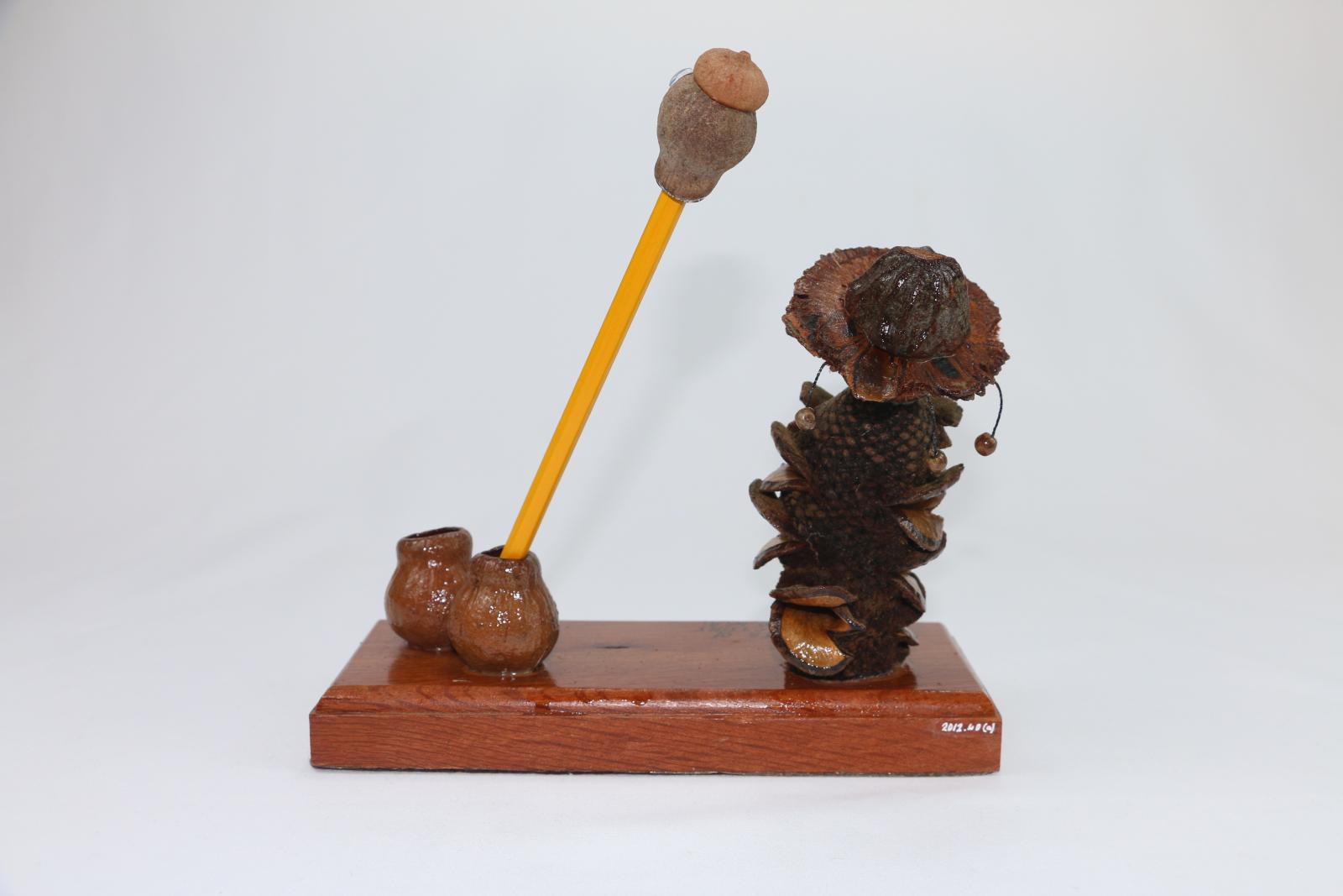 Brown, gum nut figurine sitting on a thick, rectangular wooden stand. A gum nut figure stylised as a swagman serves as an ink or pencil stand. The stylized swagman has a Banksia nut body and gumnut head with another nut as a hat with small beads hanging from it. The wooden base is lacquered with words engraved on the surface 'Compliments of/GUMNUT FACTORY/WANNEROO. W.A.' Two gum nuts on the right of the stand are glued in place and function as a pencil holder. 