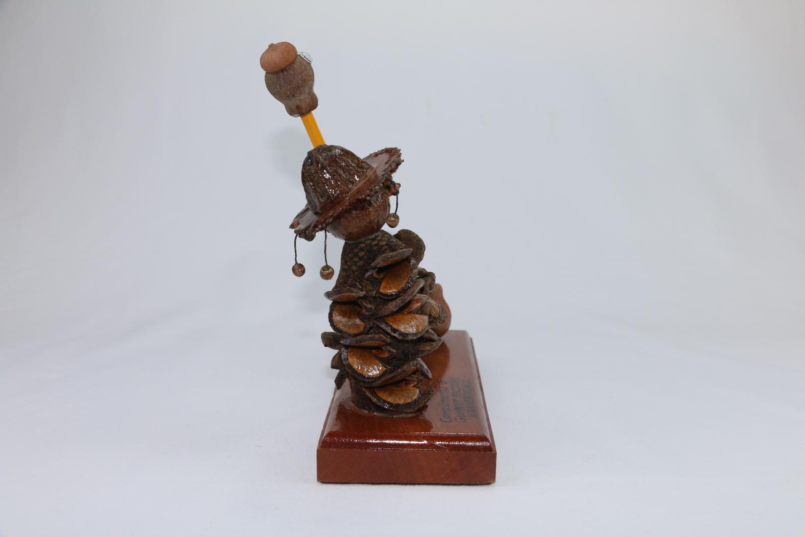 Brown, gum nut figurine sitting on a thick, rectangular wooden stand. A gum nut figure stylised as a swagman serves as an ink or pencil stand. The stylized swagman has a Banksia nut body and gumnut head with another nut as a hat with small beads hanging from it. The wooden base is lacquered with words engraved on the surface 'Compliments of/GUMNUT FACTORY/WANNEROO. W.A.' Two gum nuts on the right of the stand are glued in place and function as a pencil holder. 