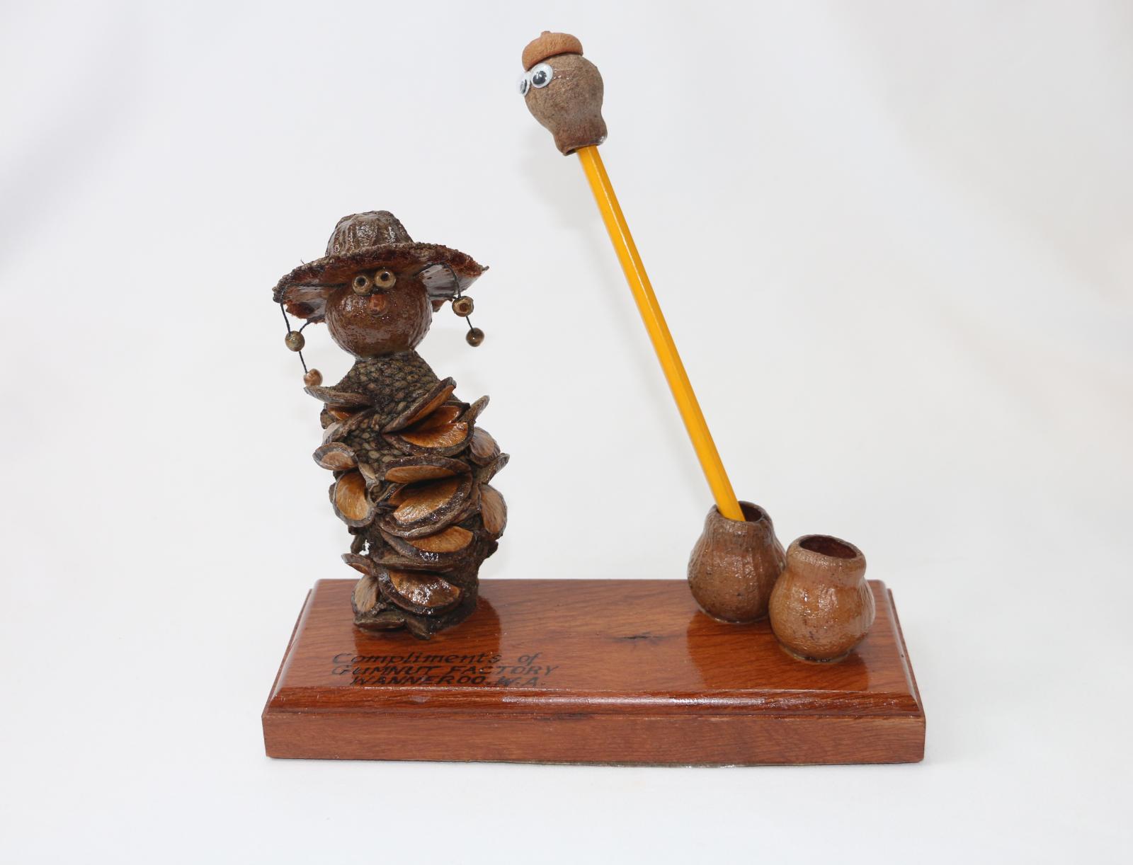 Brown, gum nut figurine sitting on a thick, rectangular wooden stand. A gum nut figure stylised as a swagman serves as an ink or pencil stand. The stylized swagman has a Banksia nut body and gumnut head with another nut as a hat with small beads hanging from it. The wooden base is lacquered with words engraved on the surface 'Compliments of/GUMNUT FACTORY/WANNEROO. W.A.' Two gum nuts on the right of the stand are glued in place and function as a pencil holder. 