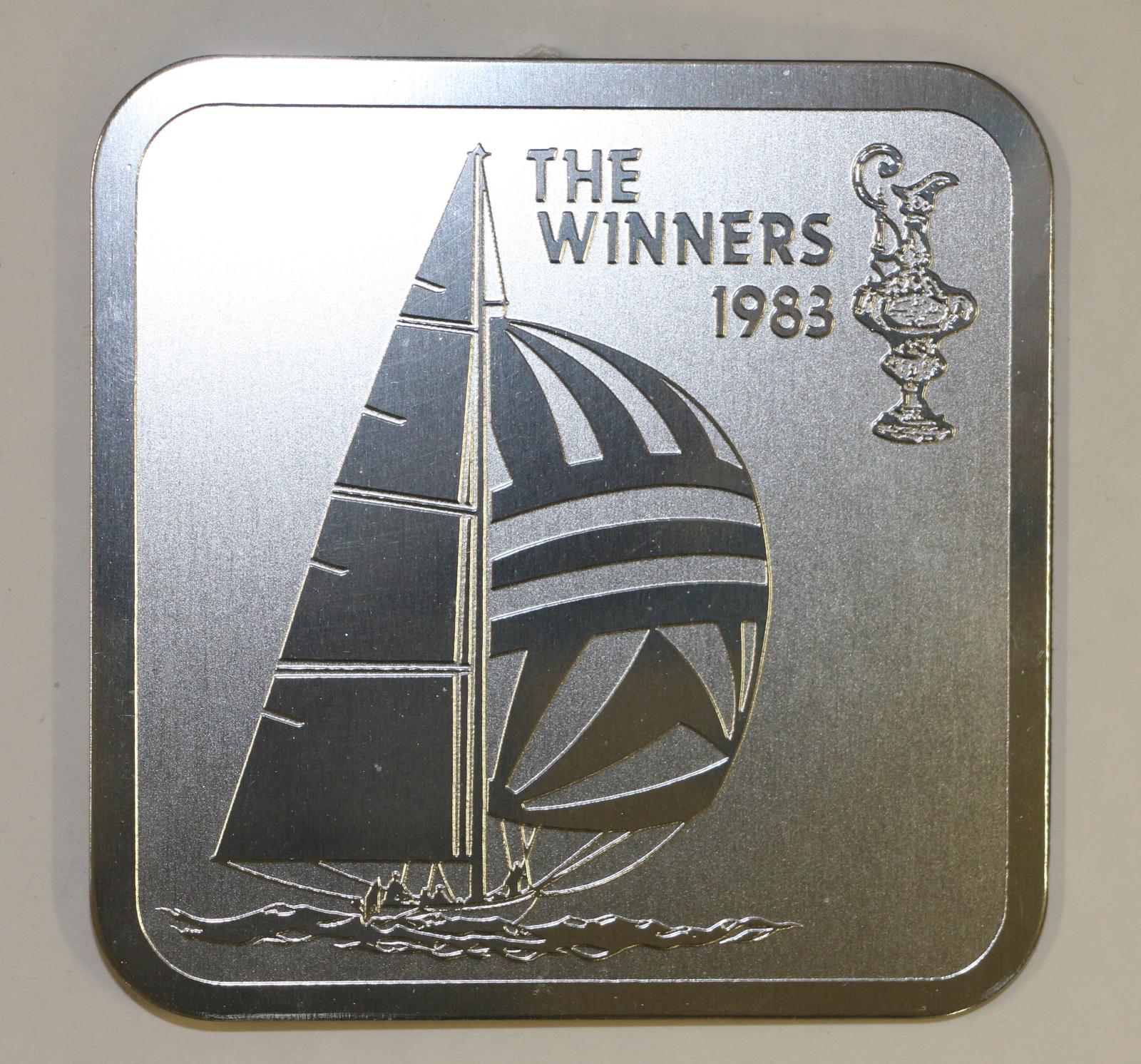 A silver coaster frame with an image of the Australia II, the America's Cup, and text stating The Winners 1983. The coaster has an embossed border. The cardboard backing has a section cut out which can be used as a stand to display the coaster upright.
