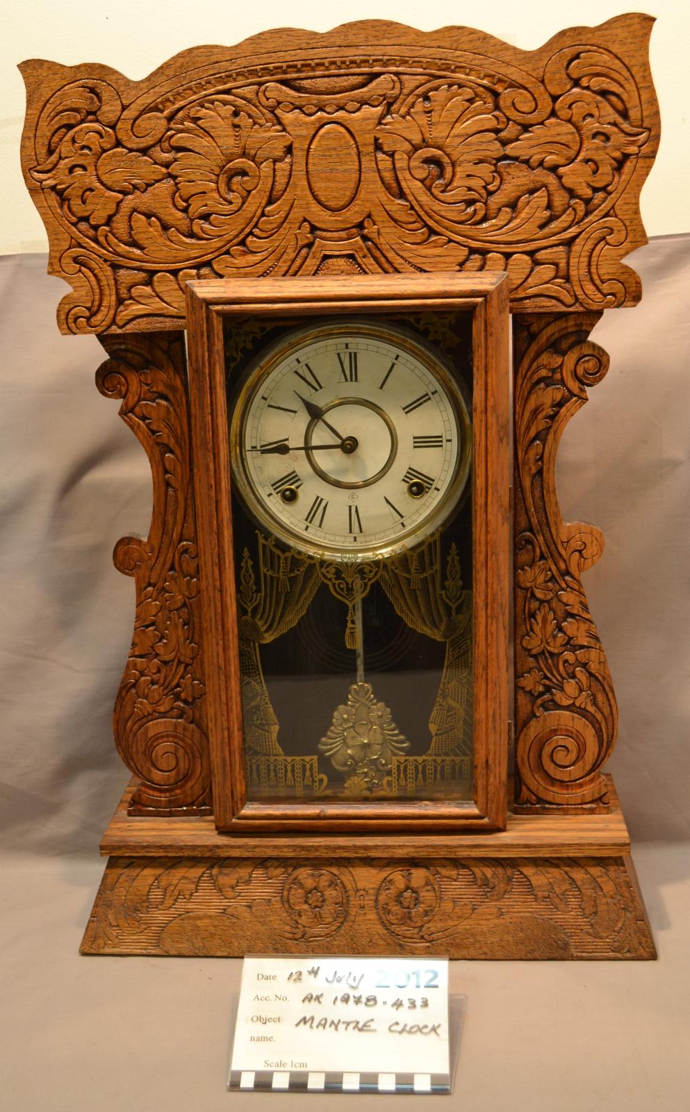 An ornate mantel clock made of wood, with the clock face and pendulum contained behind a rectangular glass door.