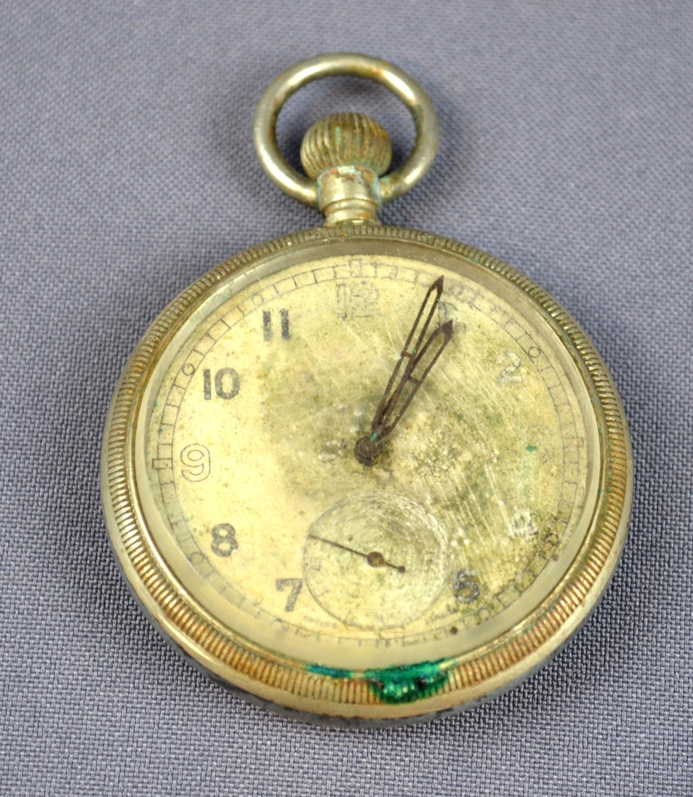 A round, gold/yellow-coloured pocket watch. The right-hand side of the face has been damaged, leaving only the numbers 7, 8, 10 and 11 still on the face. There is an unknown green mark at the 6 O'clock position of the outer rim of the watch.