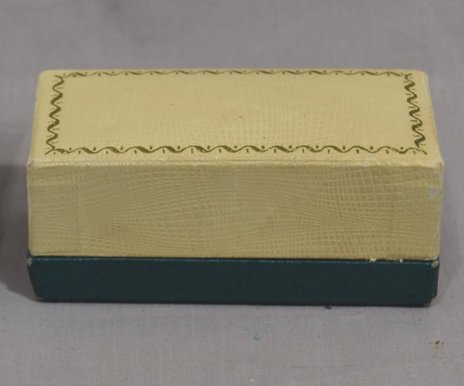 A rectangular watch box with a dark green base and cream coloured top. The top of the box lid has a gold ribbon design around it's edge.