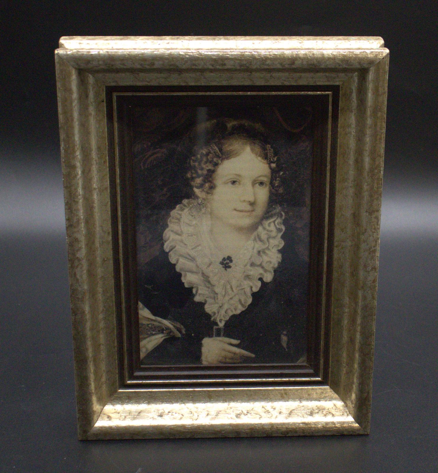 Head and shoulders picture of Mrs Henry Edward Hall, nee Sarah Theodosia Branson, on a dark mottled background. Her hair is centrally parted,  curled and piled to the back of the head.  She is wearing a ruffled collar with flower like brooch over a dark belted dress. The picture is mounted in a gold speckled frame with a brown border.