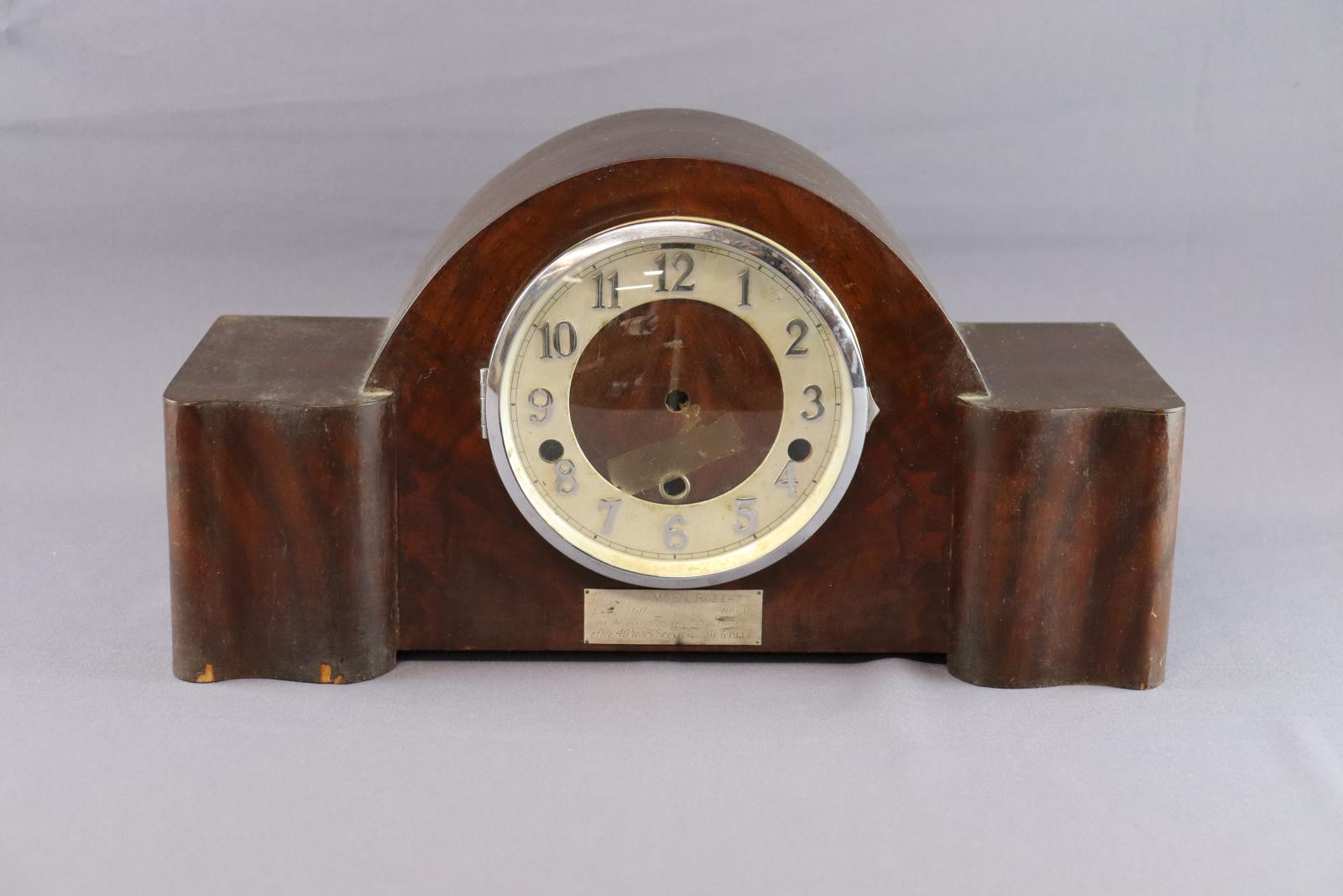 A wooden mantel clock with a rectangular base and curved top over clock face. The clock face is centered with a glass cover that opens on a hinge to the left. Metal numbers are around edge of clock face, which has no hands. There is an engraved metal plaque under the clock face.
