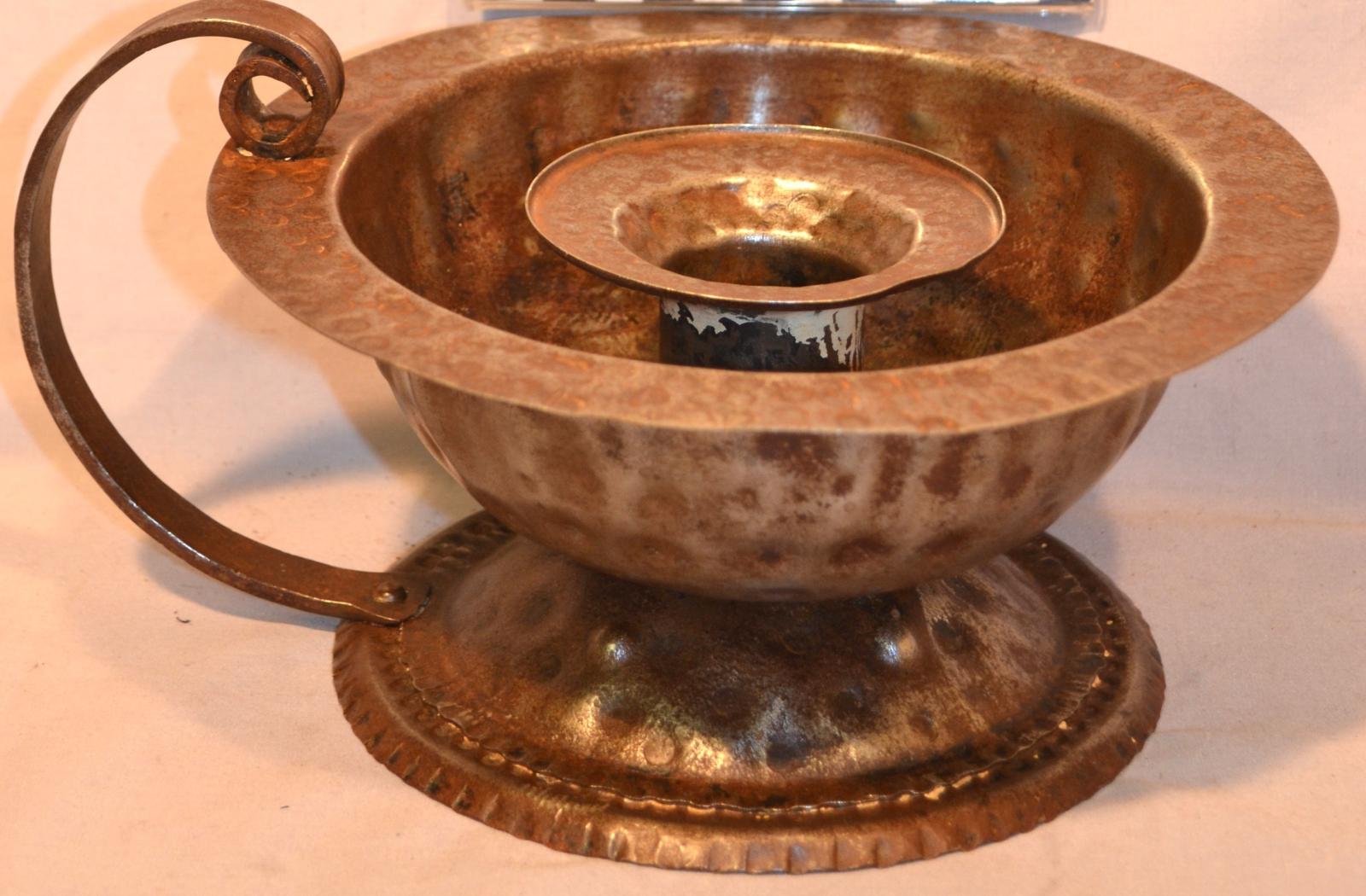 A round, copper-coloured, candle holder, made of tin. It has a bowl for the wax catchment area and a curved handle attached from the rim of the bowl to the base.