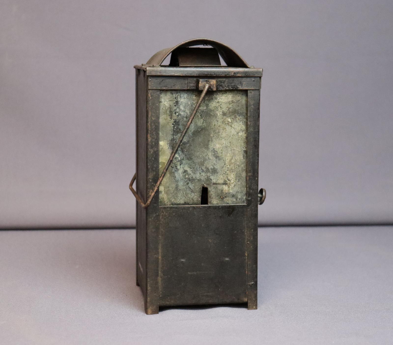 Rectangular metal case with a rectangular window taking up the top half, covered on the inside by a heavily oxidized piece of metal with a small slit at the bottom. The squared handle and burner control knob are both visible from this angle.